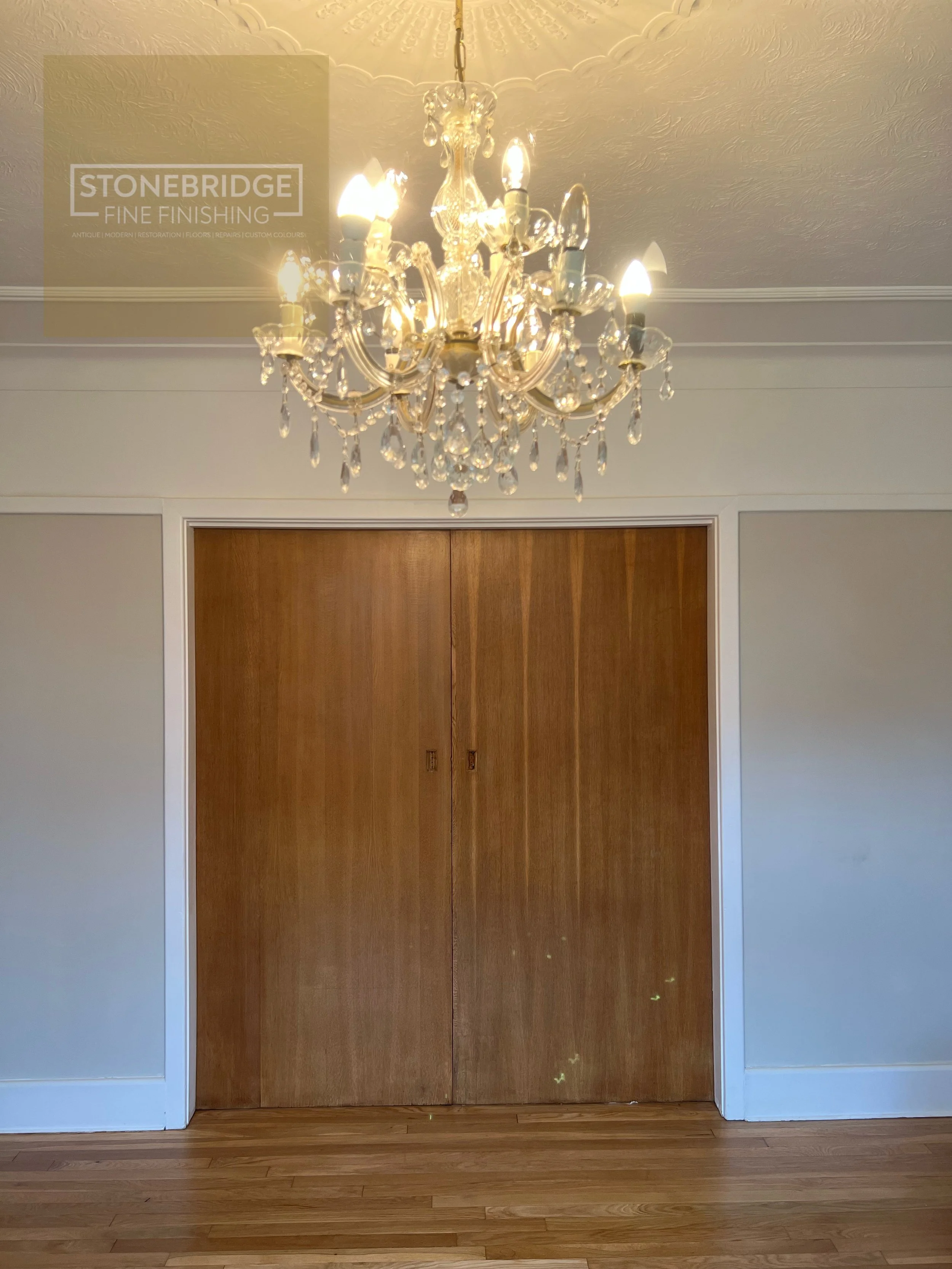 A chandelier hanging from a textured ceiling above closed wooden closet doors, with a floor of hardwood planks and white trim around the closet.