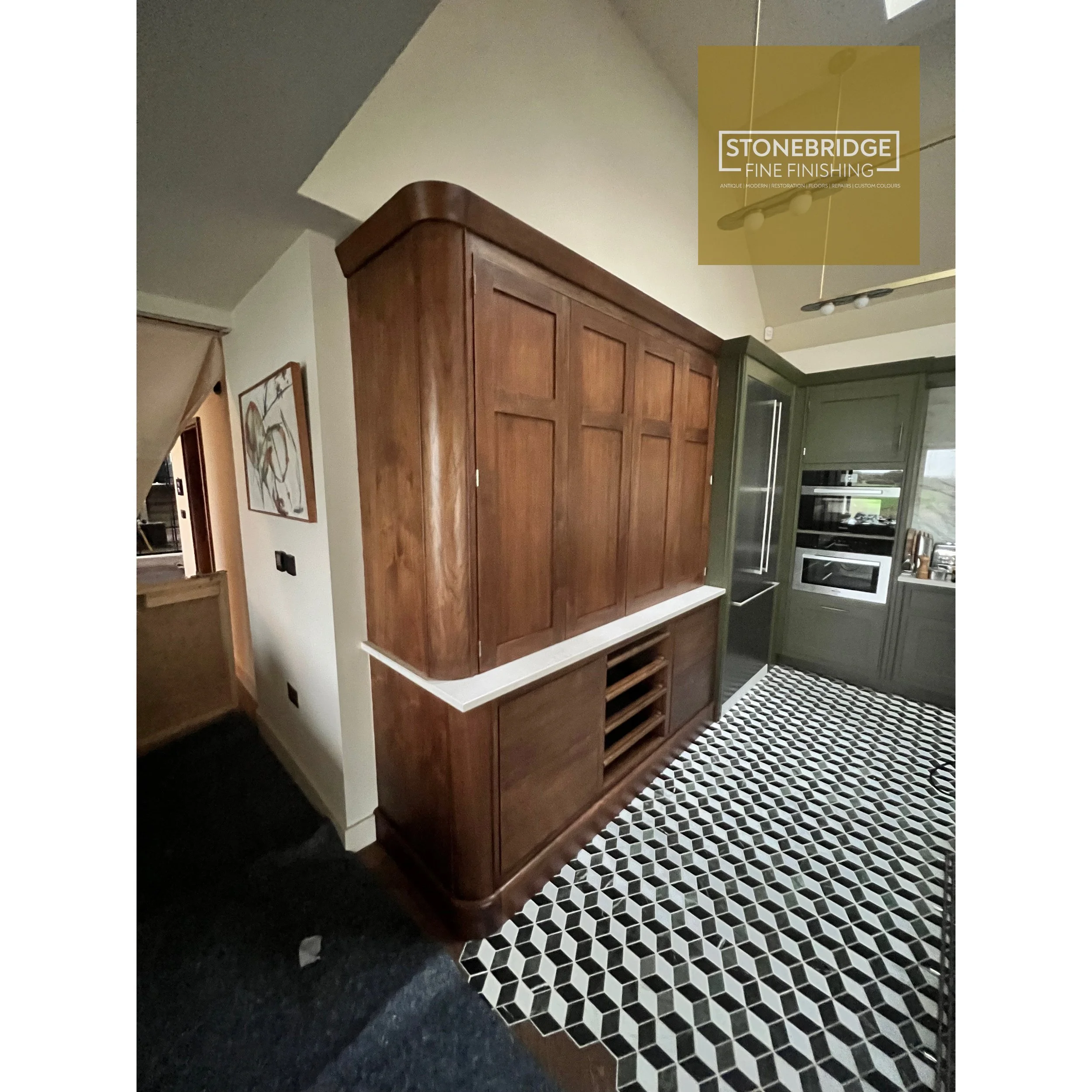 Interior view of a kitchen with wooden cabinets, a black and white patterned tile floor, and a black countertop. Part of a hallway and hanging light fixtures are visible.