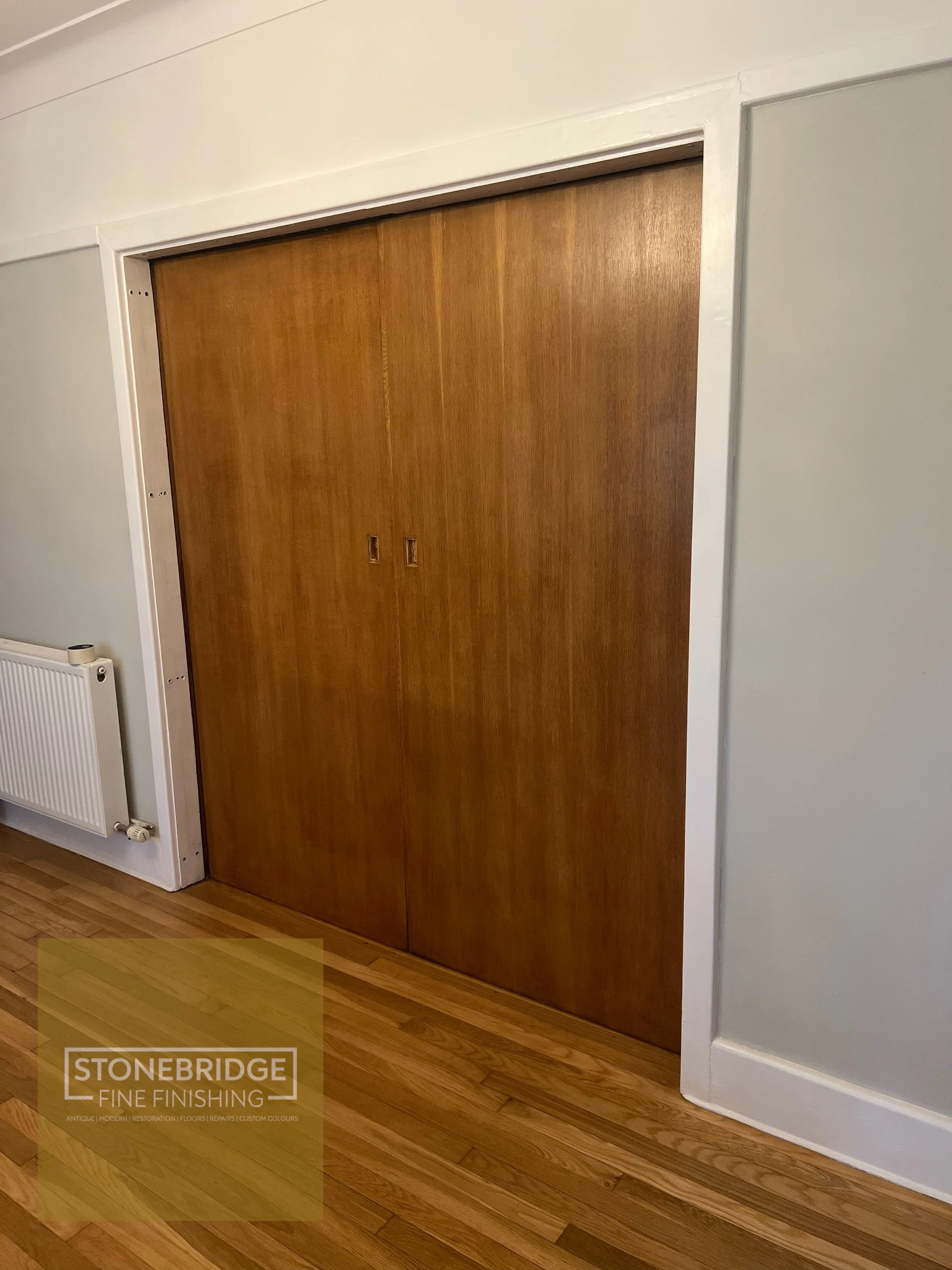 Wooden sliding door with two small rectangular handles, surrounded by white framing, next to a white wall with a radiator and wooden floor.