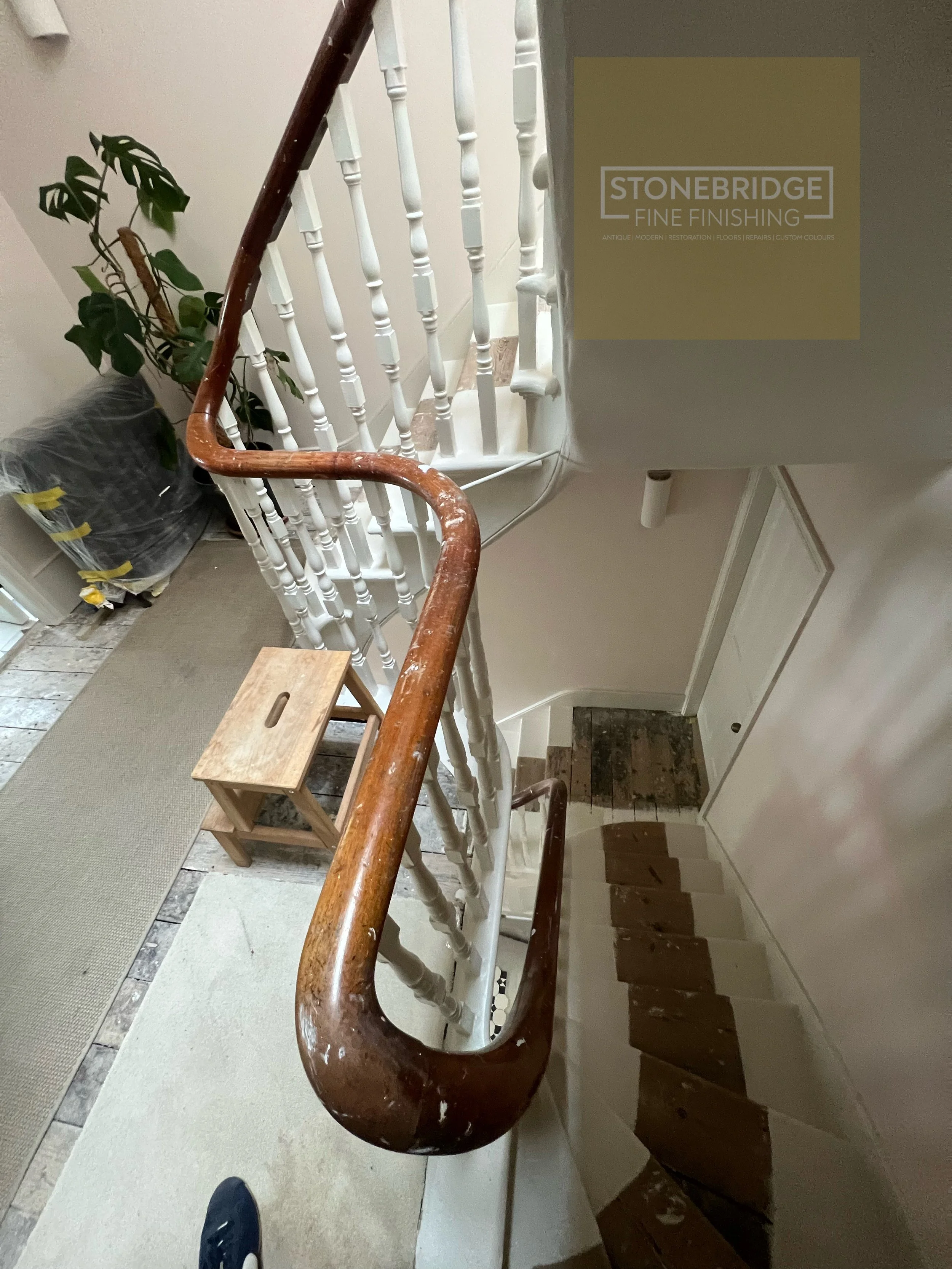 A staircase with a worn wooden handrail, white spindles, and a tiled floor at the bottom. There is a potted plant in plastic wrapping on the left side, and a beige carpet running alongside the stairs. The photo is taken from a top-down perspective, showing the stairs going down and a door at the bottom.