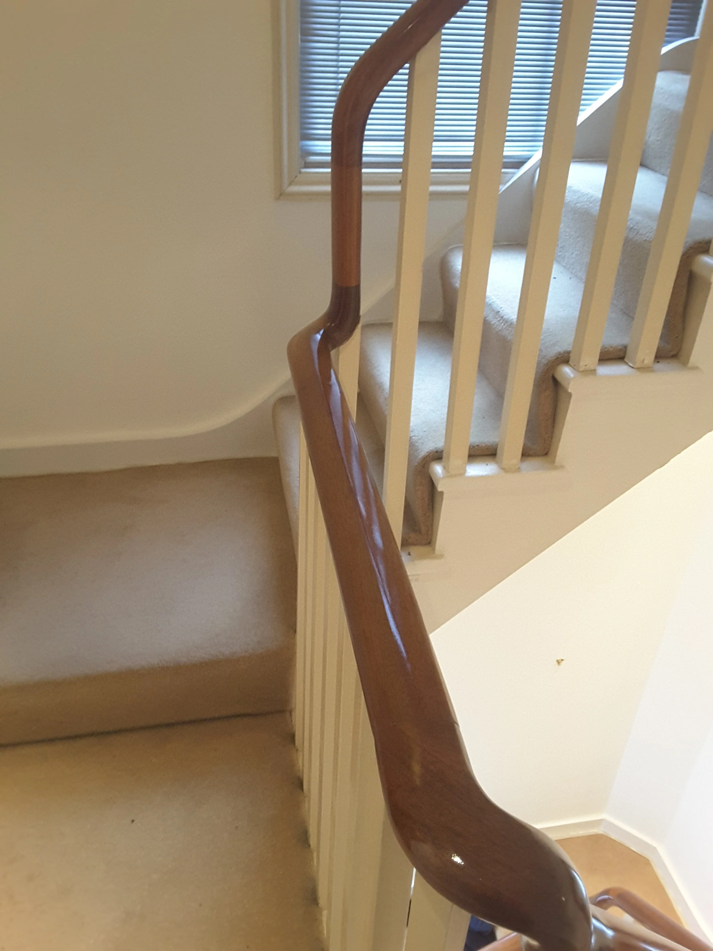 Interior view of a staircase with a wooden handrail and beige carpeting, partially obstructed by a planter or decorative item at the bottom, with a window with blinds in the background.