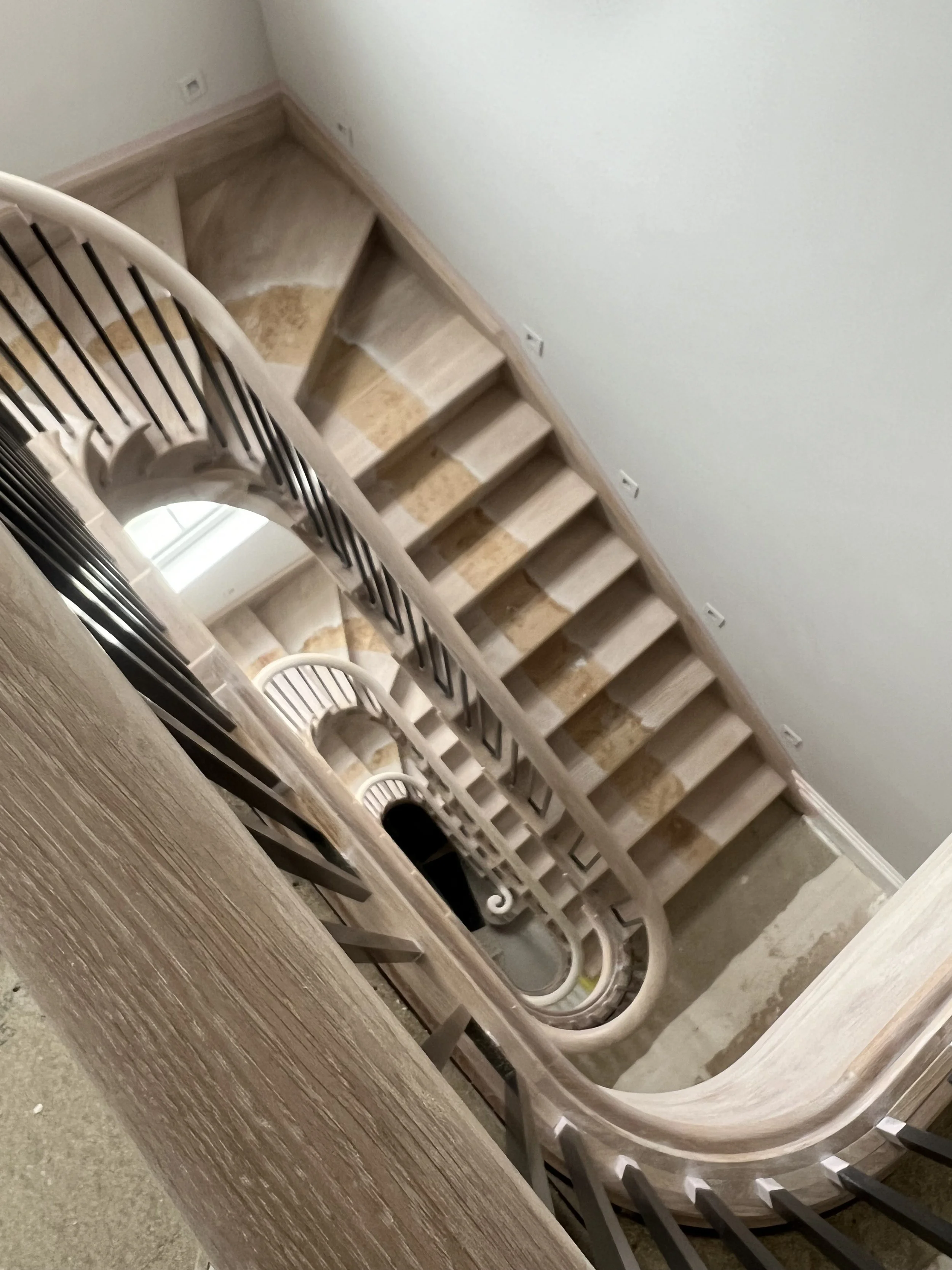 A staircase with wooden steps and black metal railings, viewed from above. The staircase curves downward around a central column, with a wall on the right side and electrical outlets on the wall.