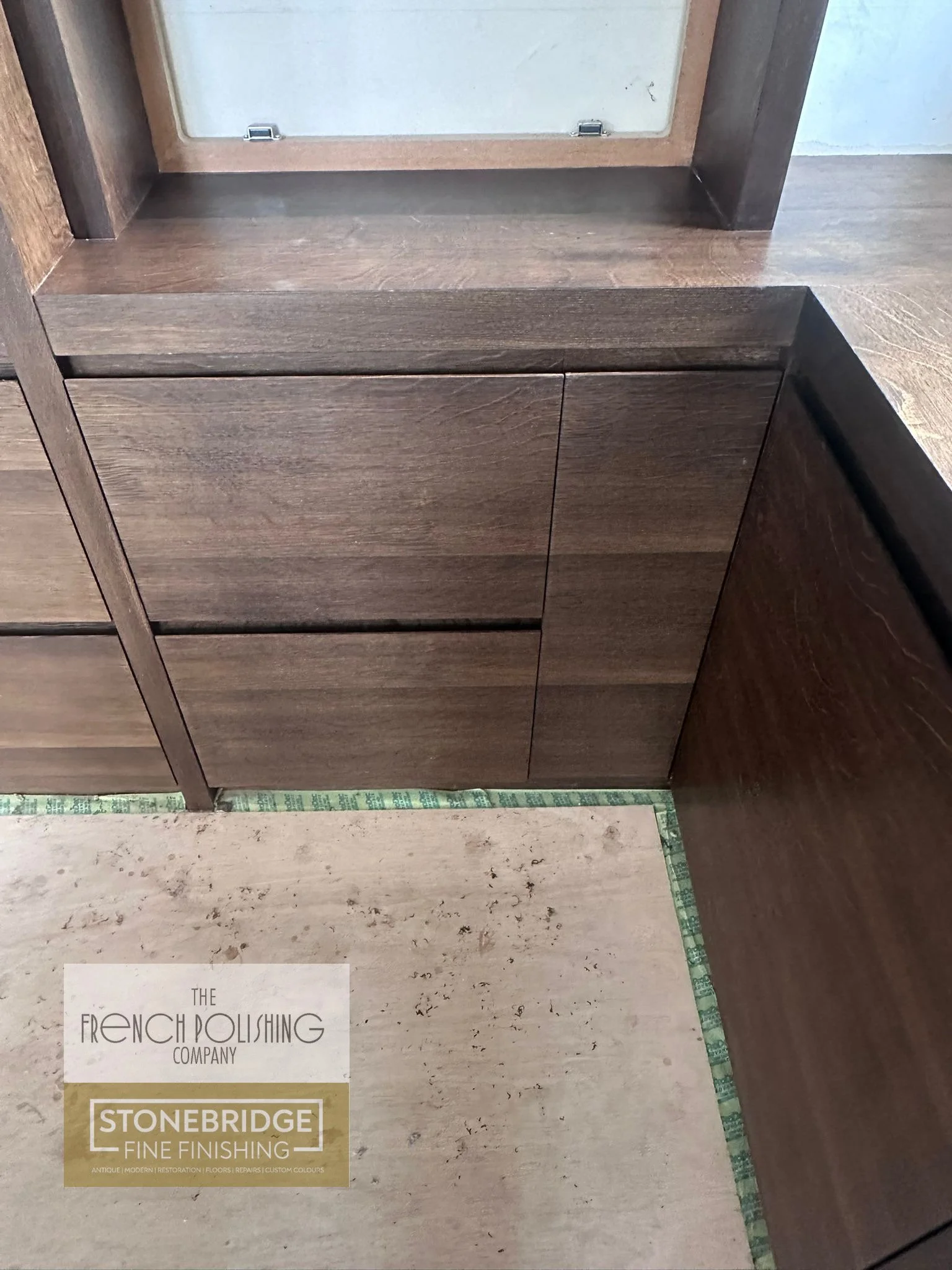 Close-up of a wooden cabinet or furniture piece with a mirror on top, with part of a sample or color swatch and a logo in the bottom left corner that reads "The French Polishing Company, Stonebridge Fine Finishing." The sample appears to be a stone o