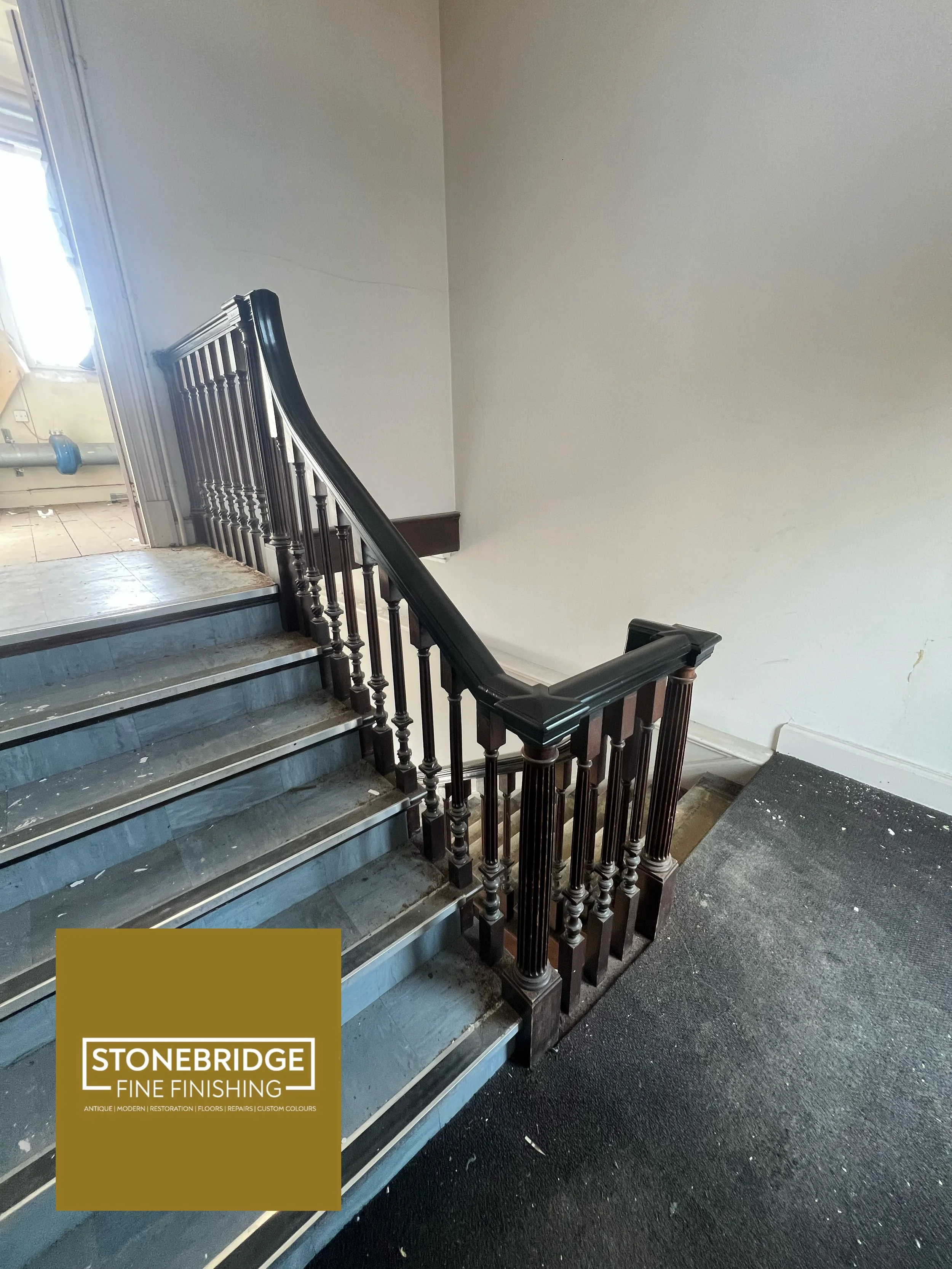 Interior of a building with a staircase featuring a dark wooden handrail and spindles, leading up to a door with a window. The wall is plain white with some scuffs. A section of black carpet is visible on the floor, with a logo for Stonebridge Fine F