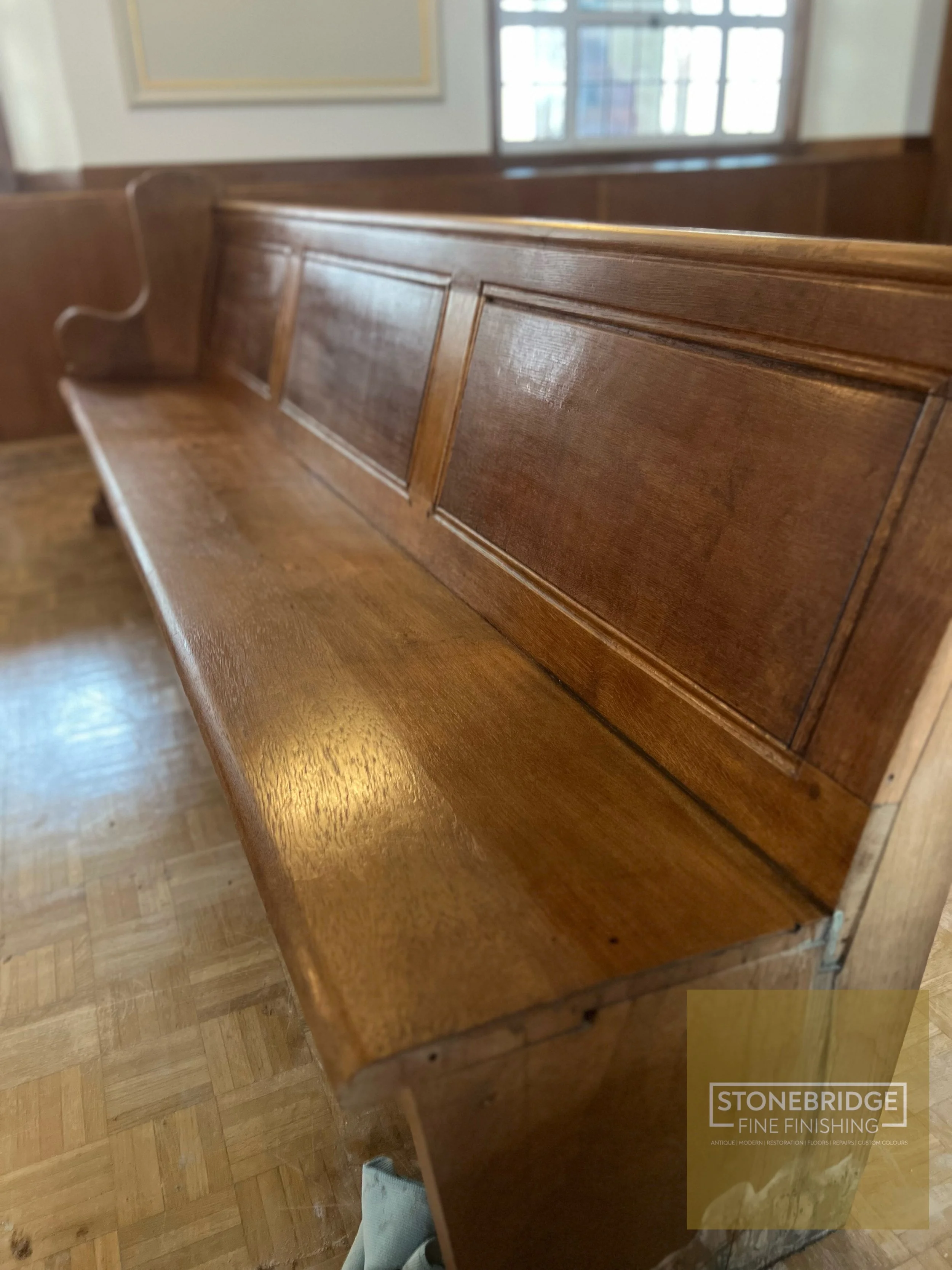 Close-up of a polished wooden church pew with detailed carving on the side, in a room with wooden flooring and large windows in the background.