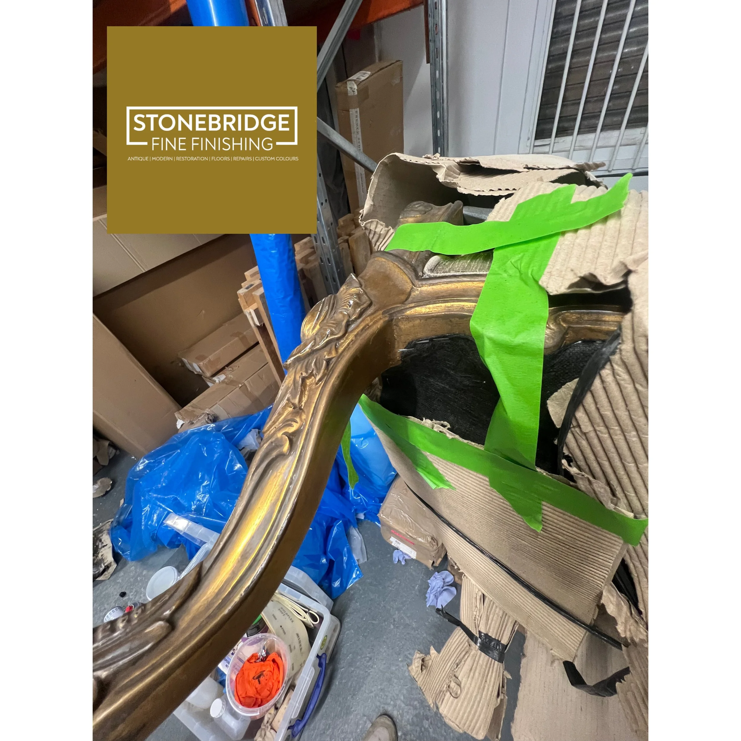 Workshop or storage space with shelves filled with boxes, paint supplies, and miscellaneous items. Bright green tape and crumpled paper are visible on the floor, and a cardboard box with a sculpture or decorative object is in the foreground.