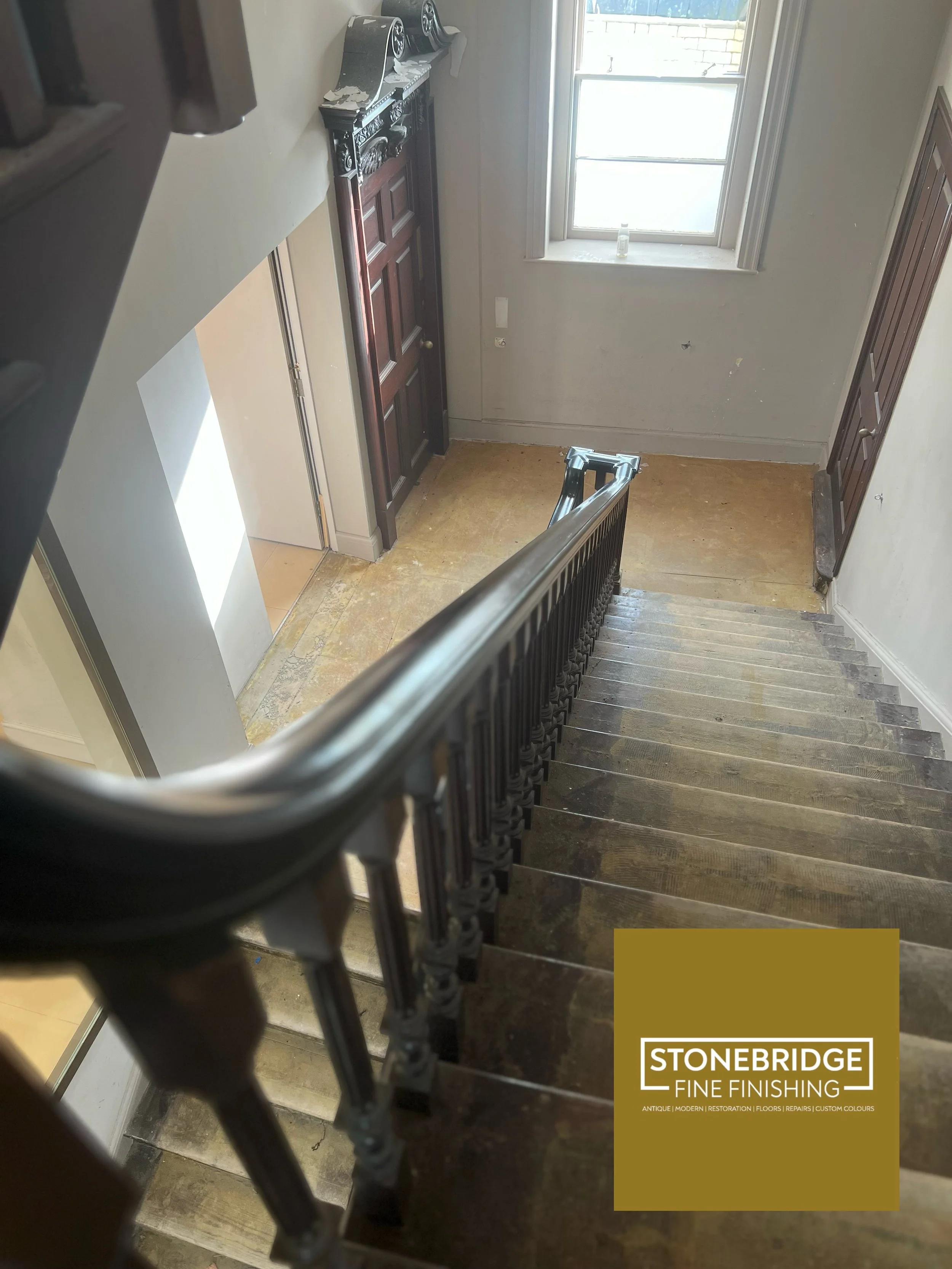 View of a staircase from the top, looking down, with a wooden handrail and a staircase with wooden floorboards. At the bottom, there is a window with a bottle placed on the window sill. To the left, part of a door is visible, and to the right, there 