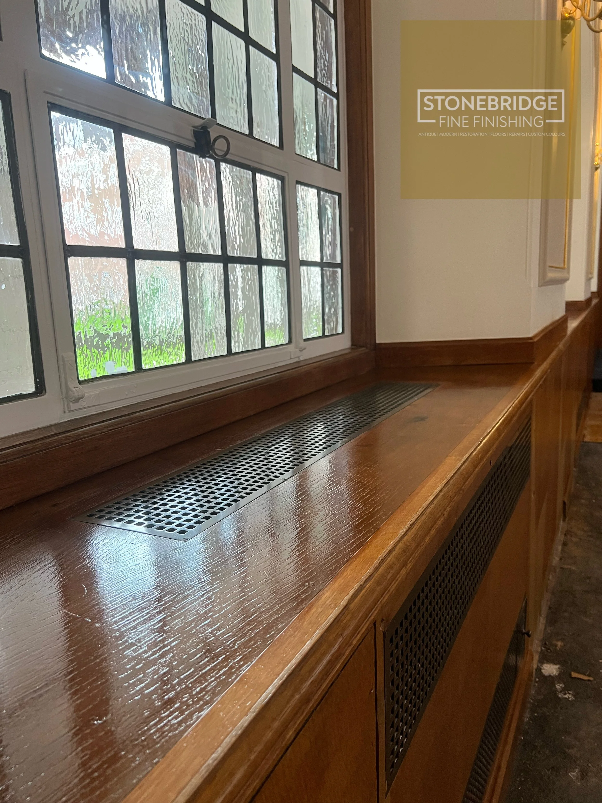 Interior view of a wooden window ledge with a decorative grate and a large window with a black metal handle. The window has smudges and condensation, and the background shows a rainy scene outside.