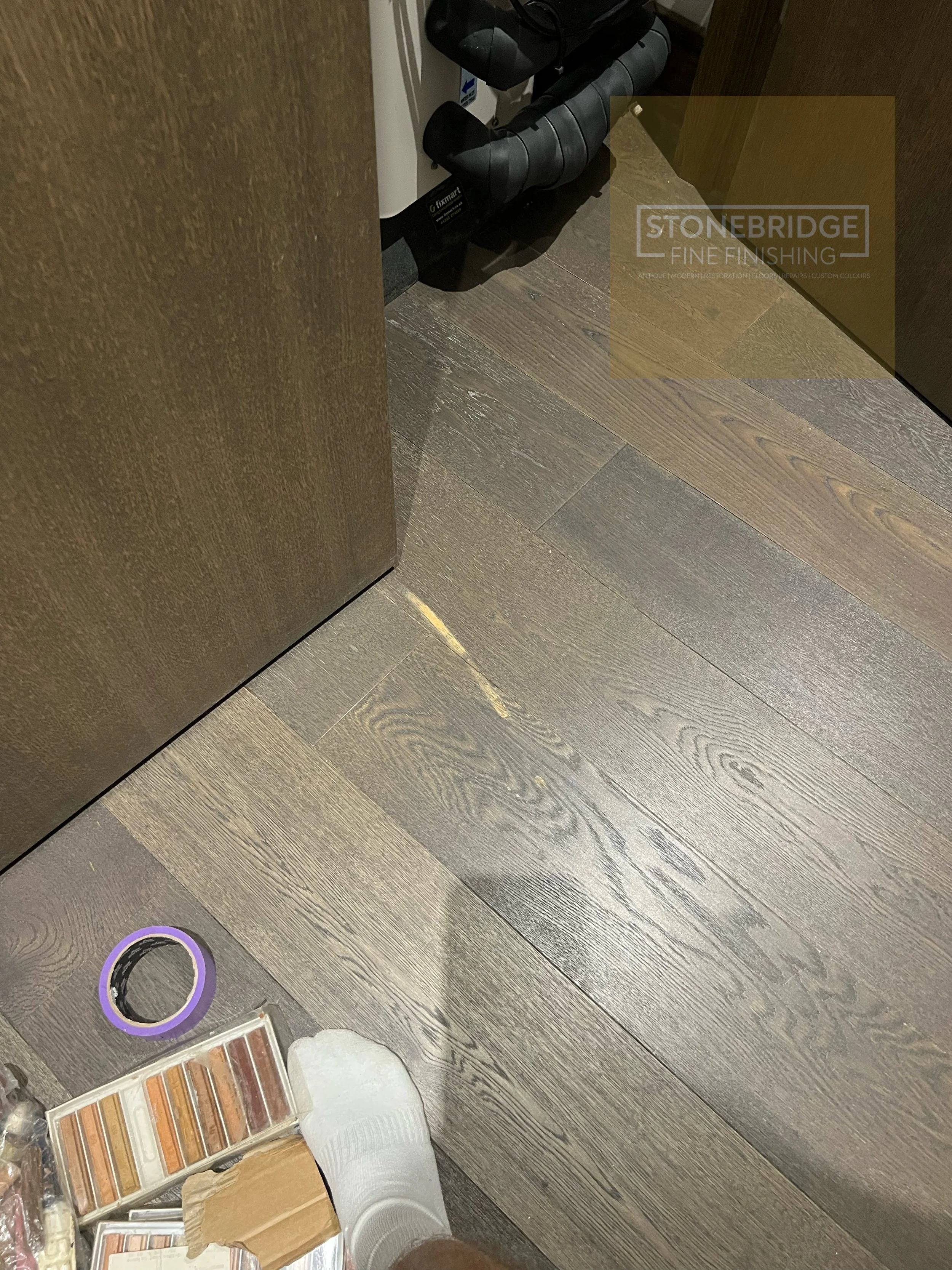 Interior of a room with dark wood flooring, a partially visible brown door, and various paint and flooring samples on a table and floor. There is a roll of painter's tape, a color palette, and a person’s feet wearing white shoes visible in the bottom of the image.