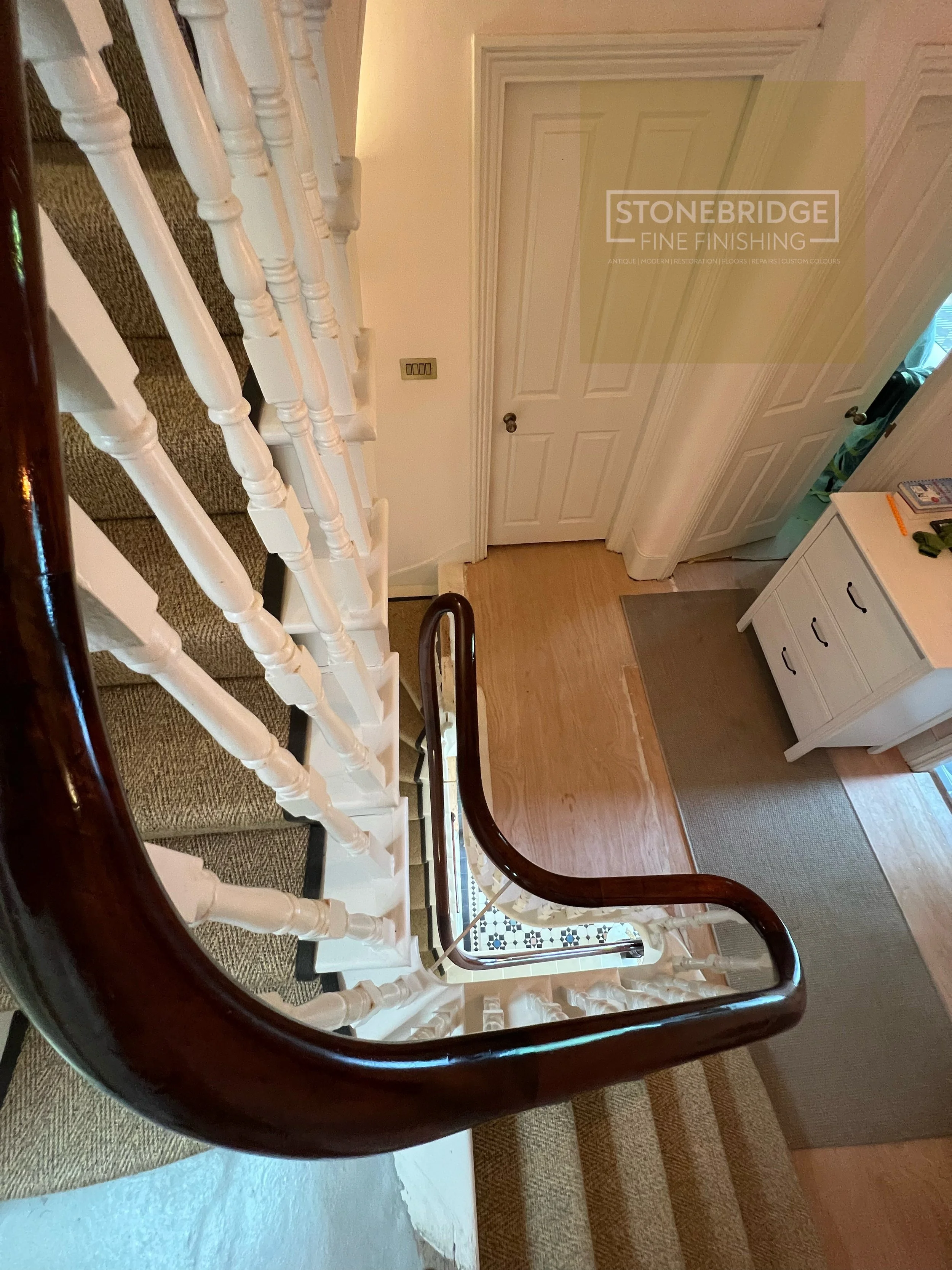 View of a staircase with white spindles and a dark wooden handrail, looking down from the top, showing a doorway at the bottom of the stairs and a hallway nearby.