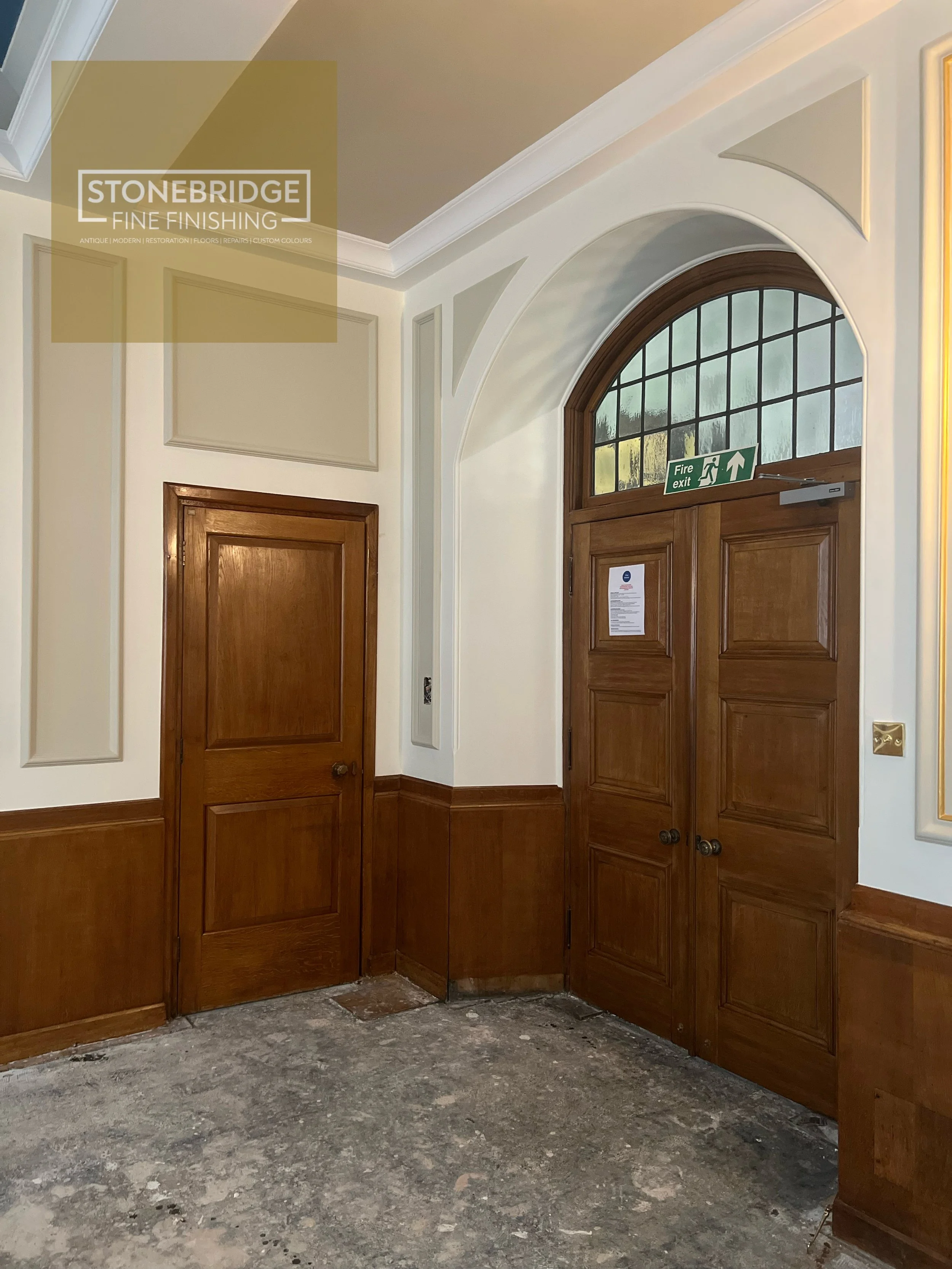 An interior room with wooden paneling and doors, with a large arched window above, and an LED exit sign. The floor appears to be under renovation or unfinished.