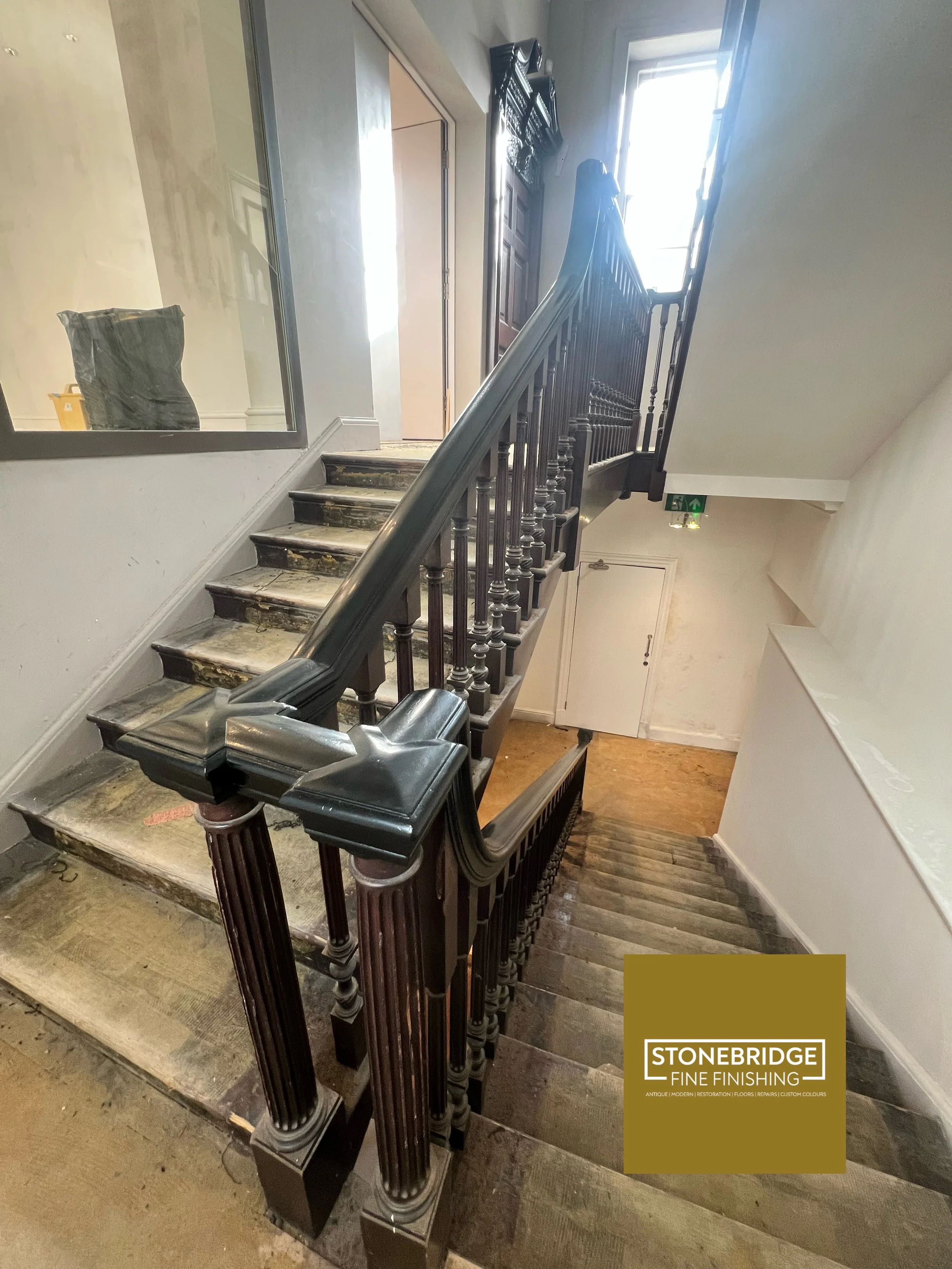 Interior of a building staircase with a black ornate handrail and decorative columns, showing worn steps and a window at the top allowing natural light in.