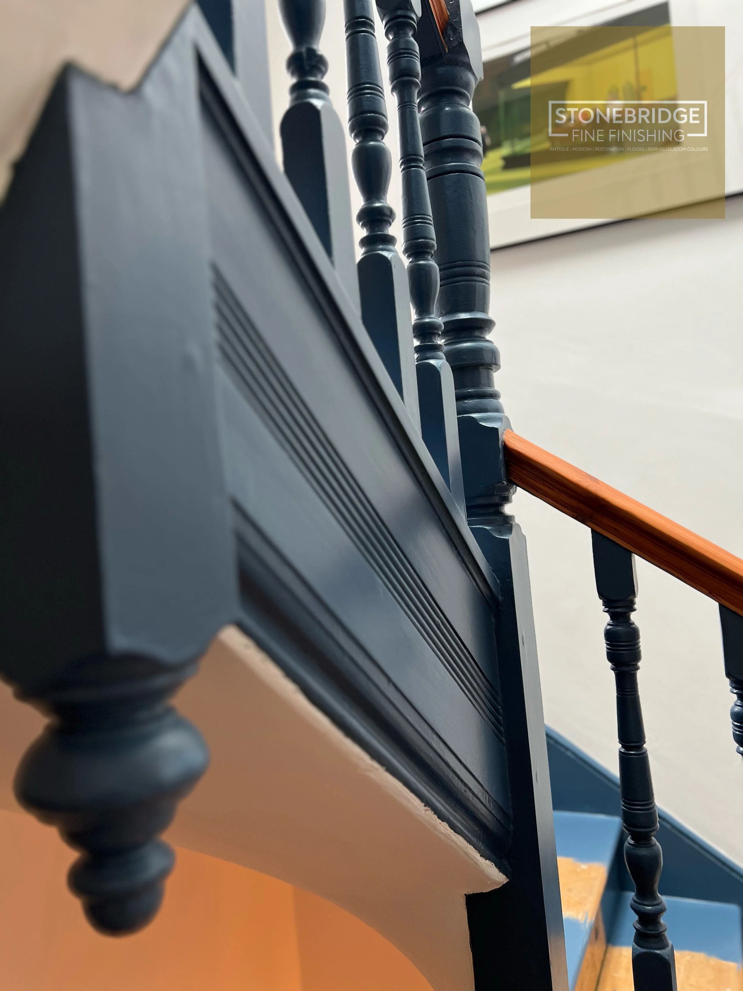 Close-up of a staircase with black wooden spindles, a wooden handrail, and framed artwork on a white wall in the background.