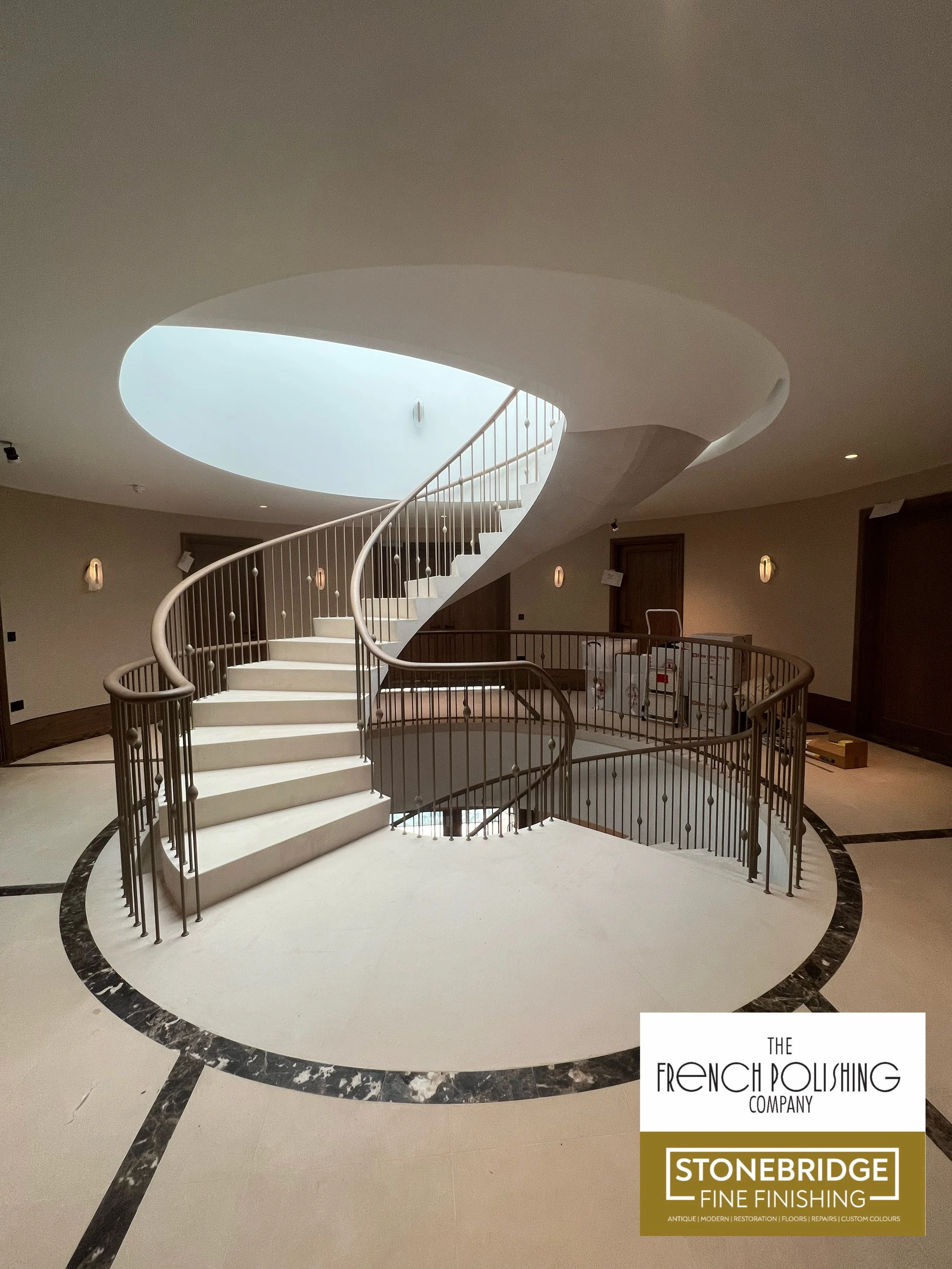 An interior view of a building with a spiral staircase, a circular skylight, and beige walls. There are wall-mounted lights and some boxes and equipment on the floor. A logo in the bottom right corner reads "The French Polishing Company, Stonebridge 