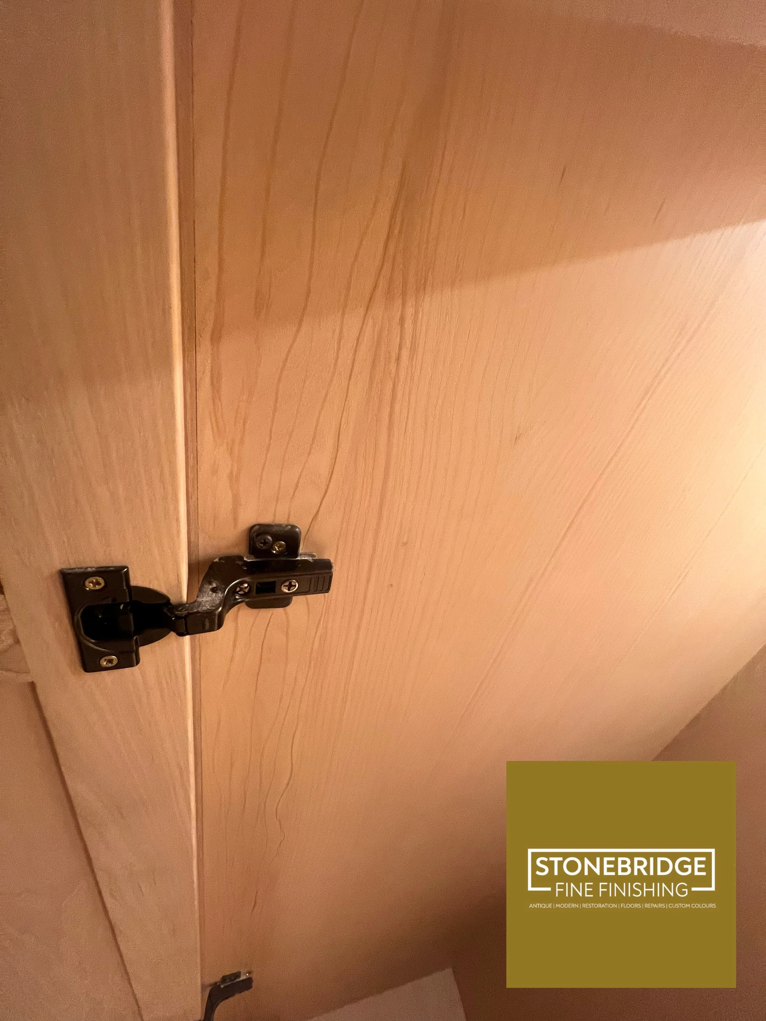 Close-up of a wooden cabinet door with a black hinge and a Stonebridge Fine Finishing logo in the lower right corner.