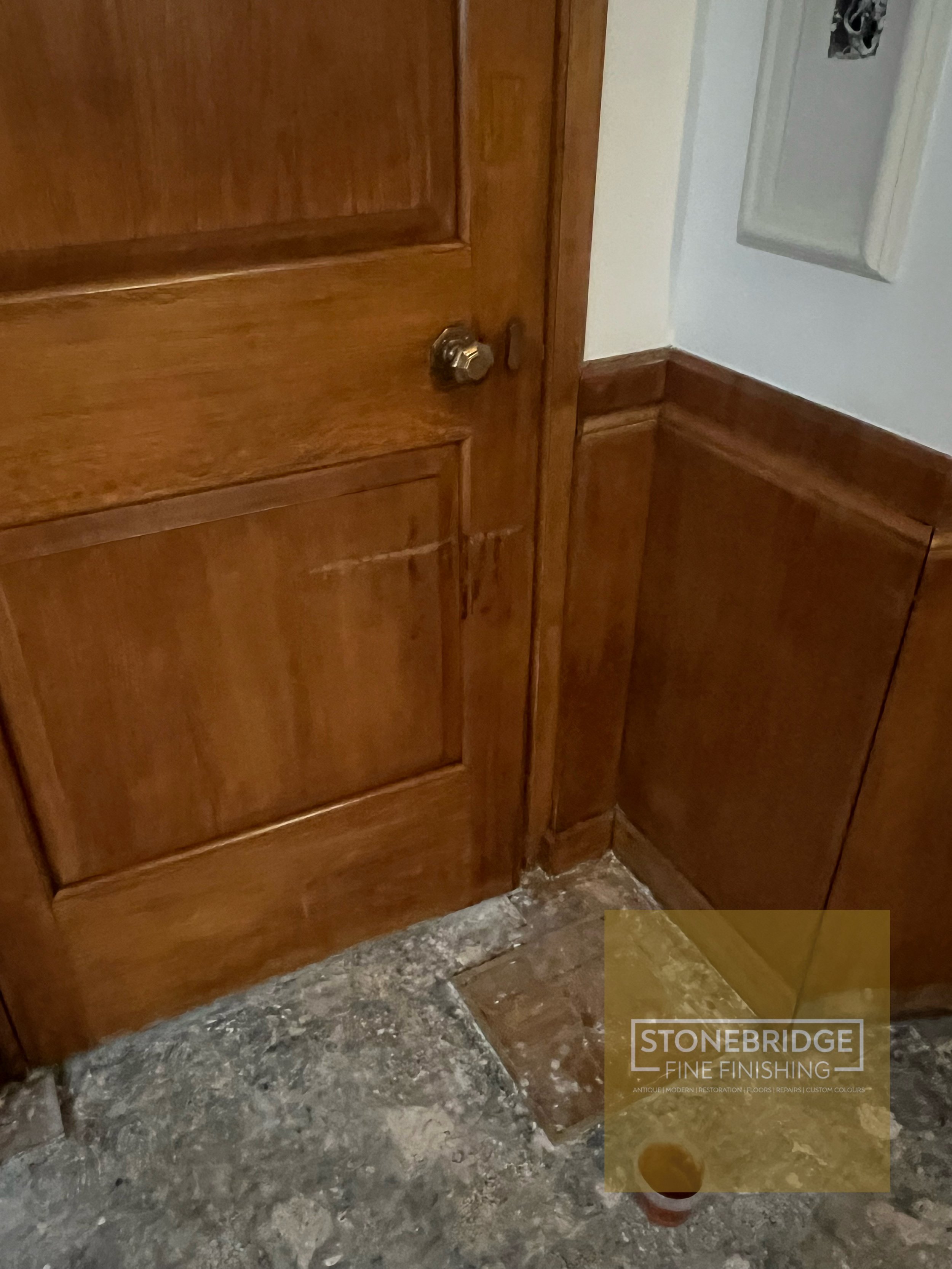 Close-up of a wooden cabinet or door frame with a metallic door knob, next to a white wall and electrical panel, with a marble or stone floor and a small cup or container on the floor.