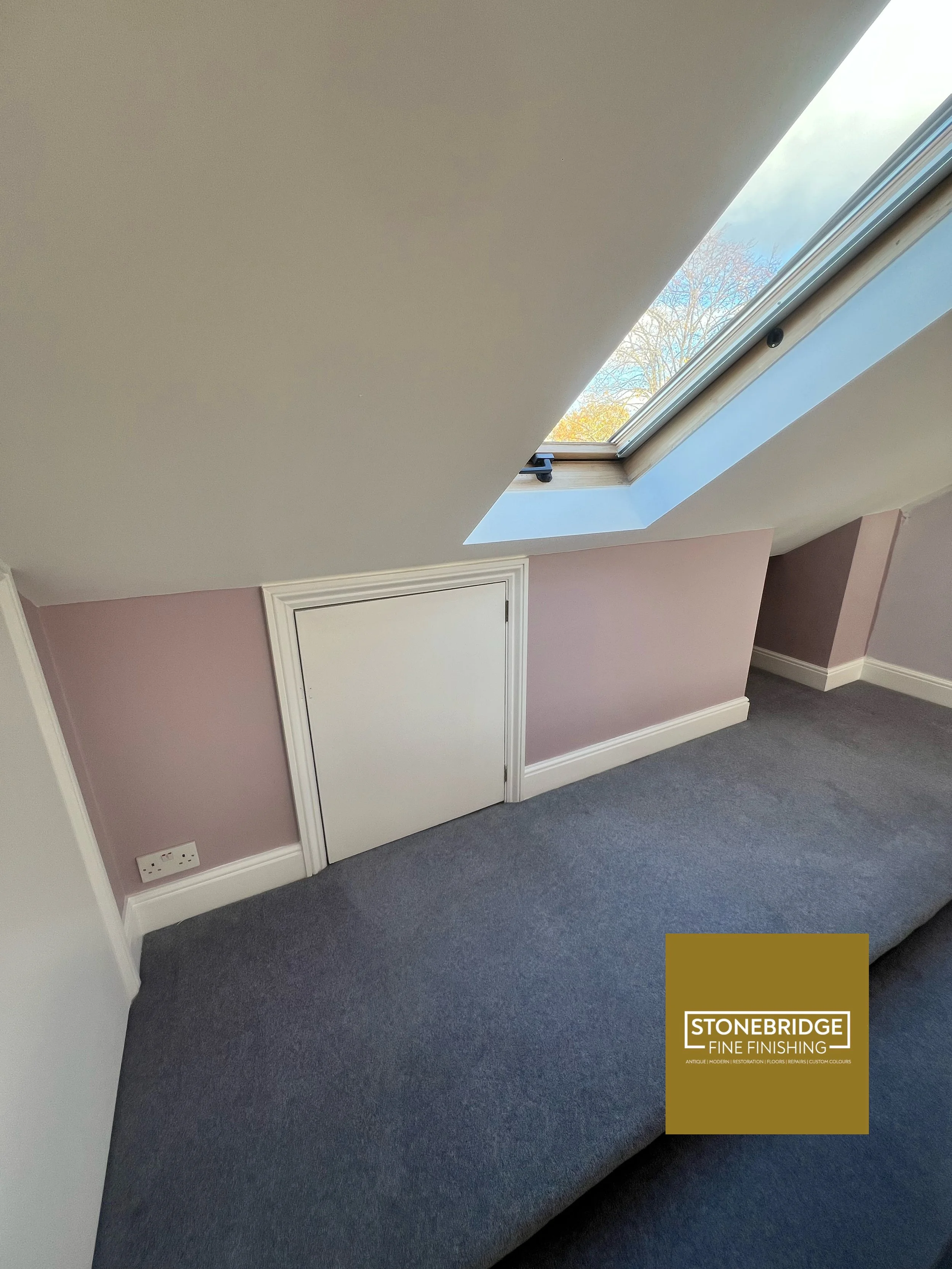 A room with a sloped ceiling and large skylight window, pink walls, a closed white door, and grey carpeted flooring, with a Stonebridge Fine Finishing logo in the bottom right corner.