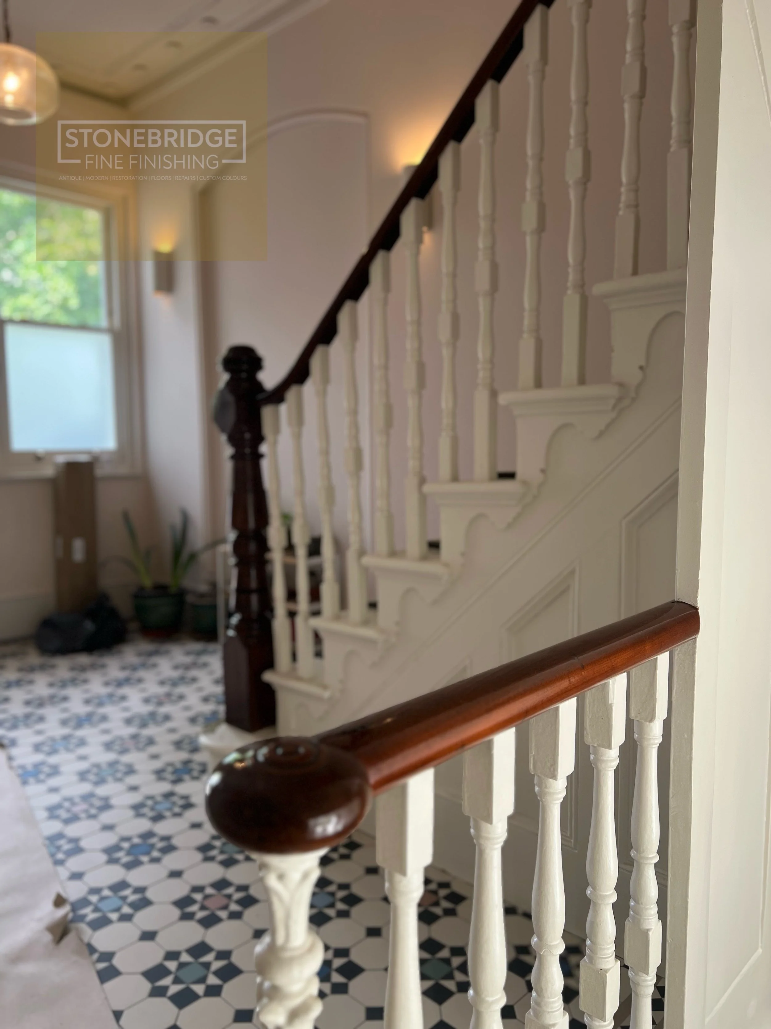 Interior view of a staircase with white balusters and a wooden handrail, black and white patterned tile floor, window with natural light, plants, and warm wall lighting.