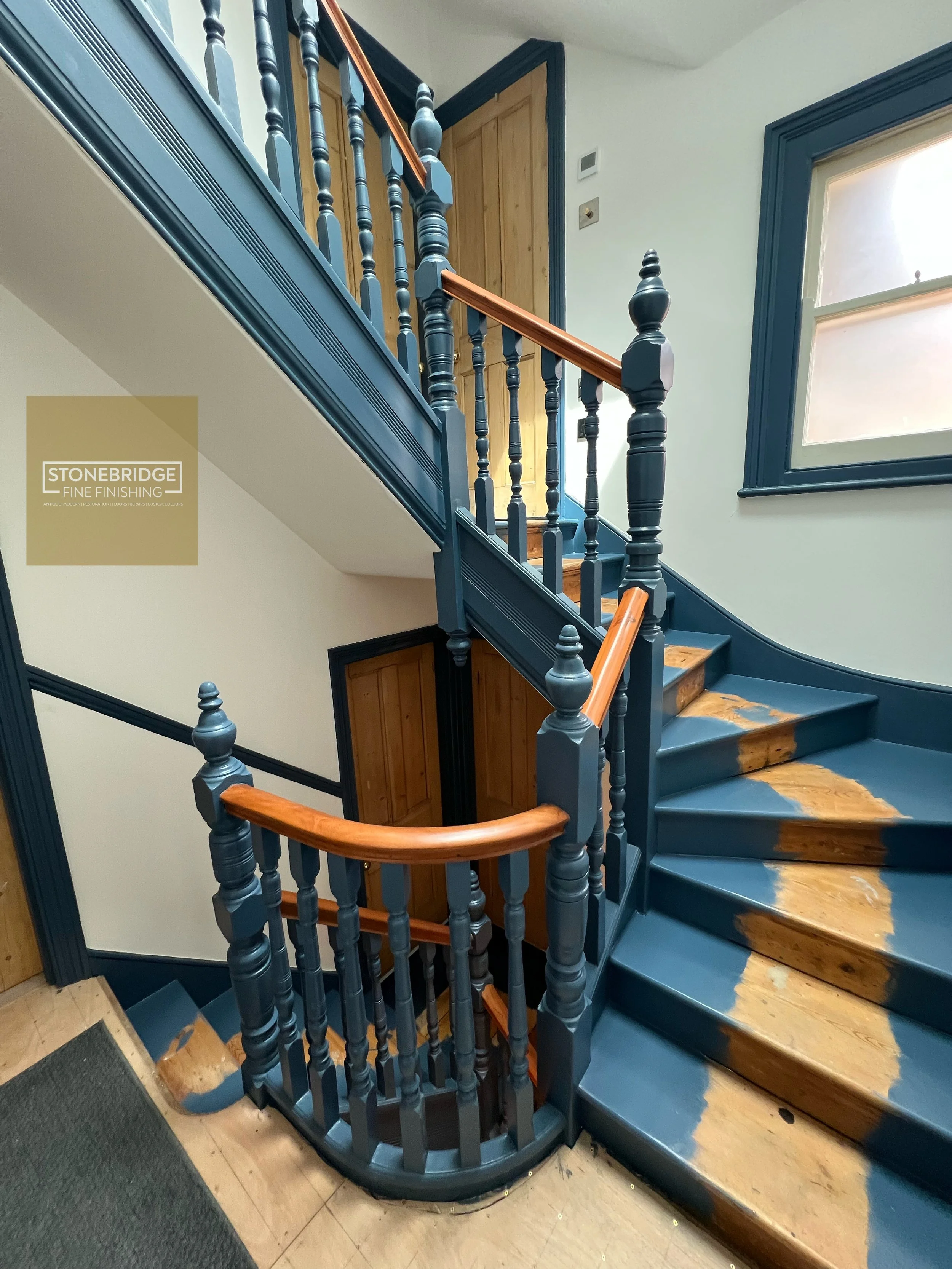 Interior of a staircase with dark blue railing, wooden balusters, and steps with a combination of natural wood and blue painted surfaces. There is a window with blue trim on the wall to the right.
