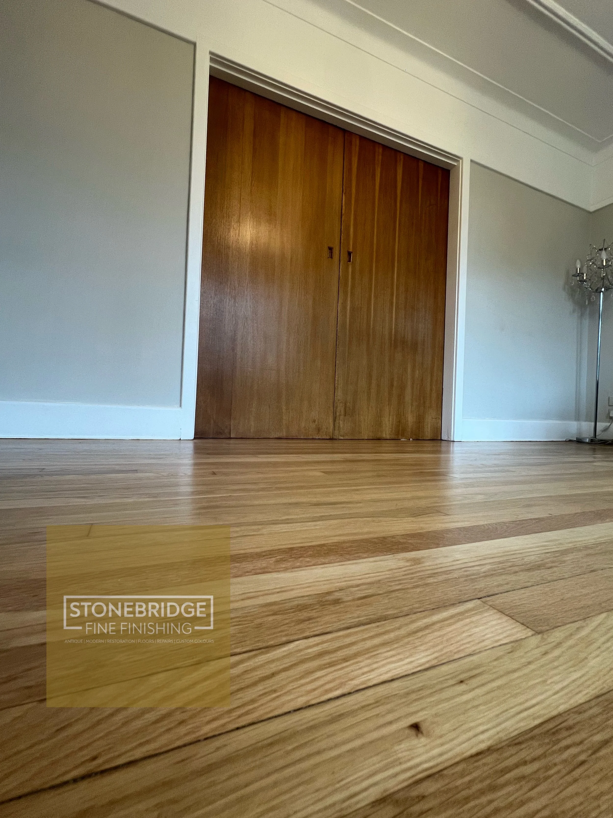 A wooden closet door in a room with white walls and light wood flooring. There is a logo in the bottom left corner that reads 'STONEBRIDGE FINE FINISHING' with additional text about their services.