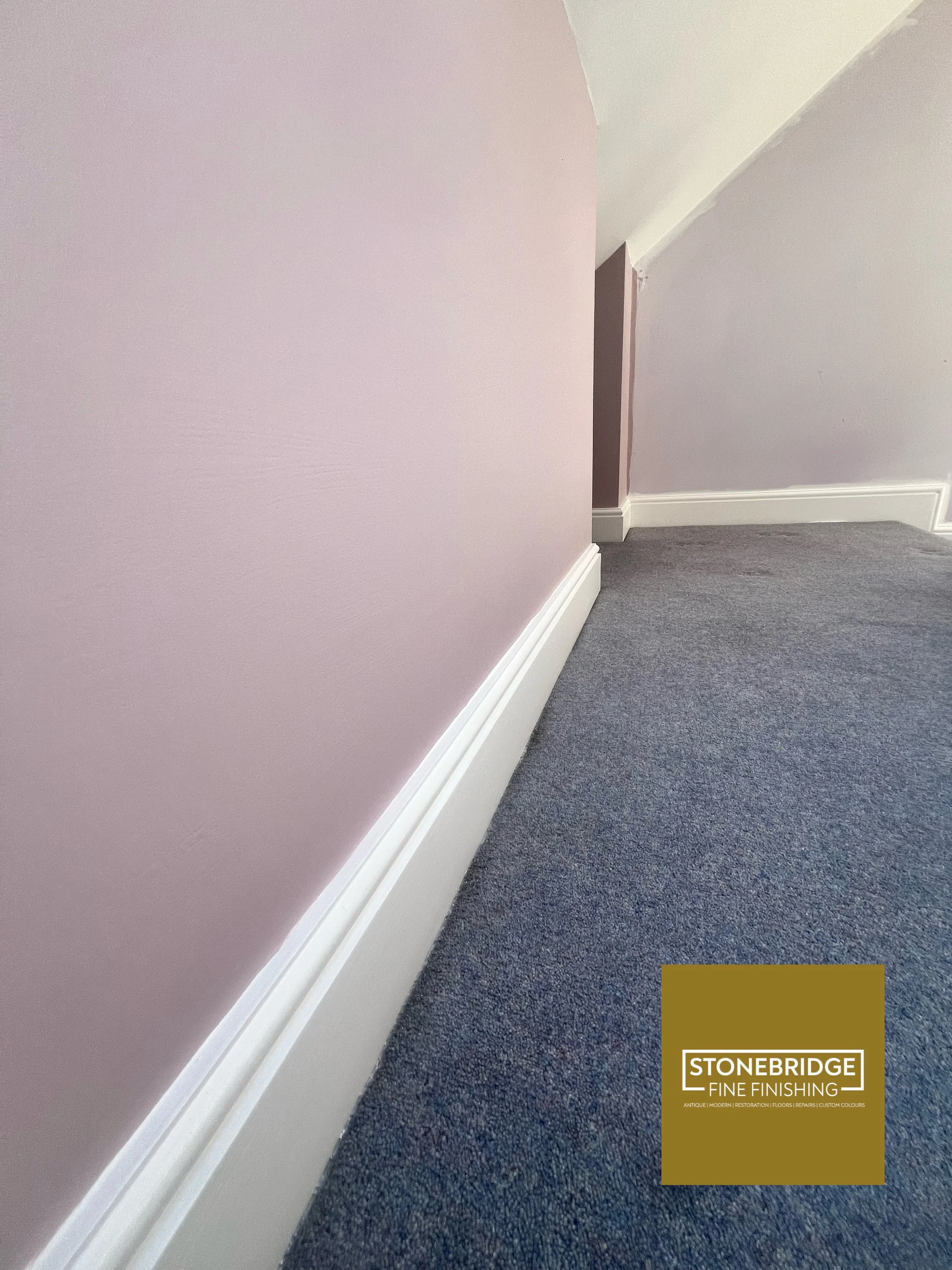 Interior corner of a room with a pink wall, white baseboard, purple wall, and gray carpeted floor, featuring a logo for Stonebridge Fine Finishing.