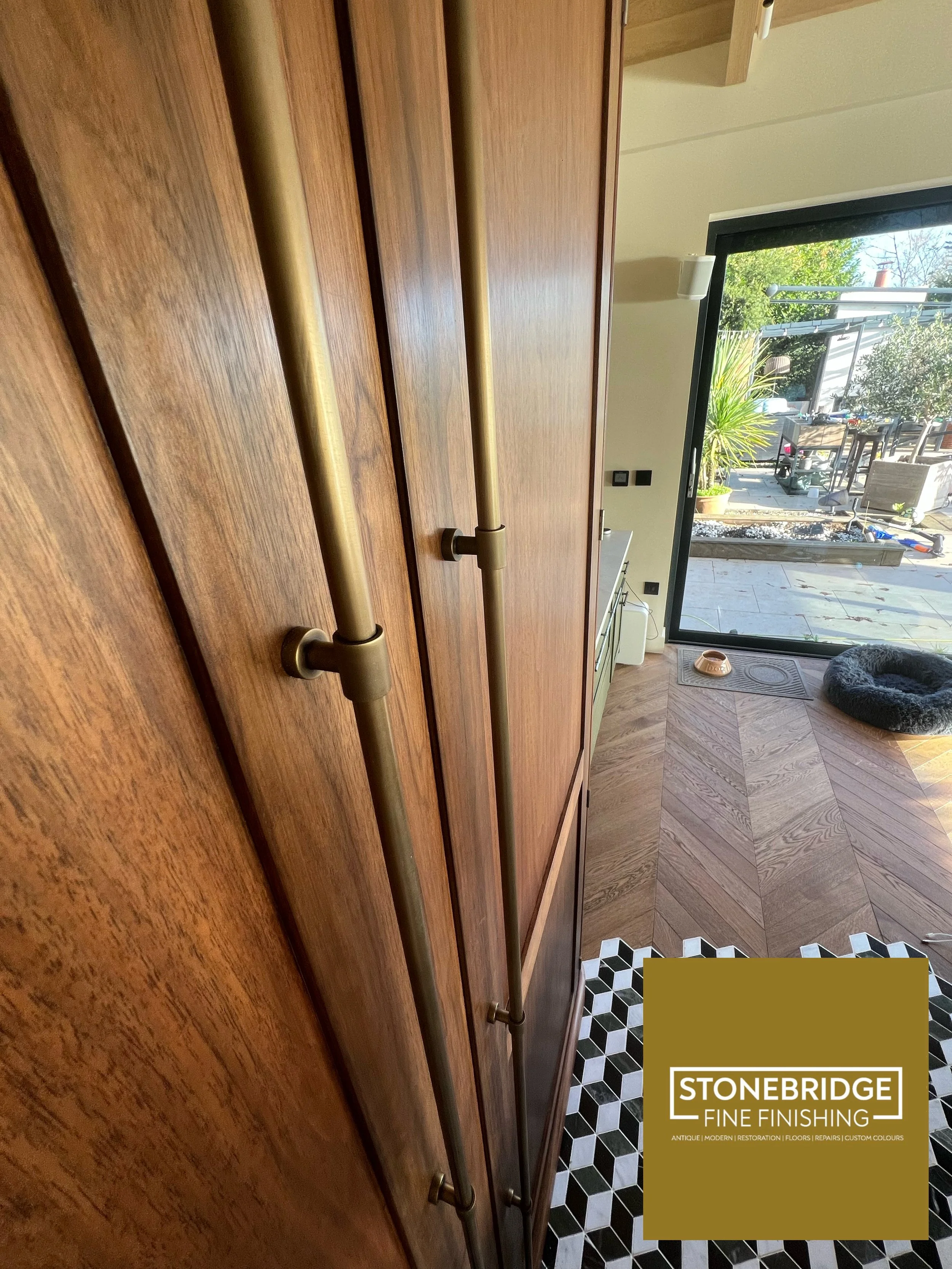 A wooden cabinet with two brass rods mounted vertically, a black and white geometric patterned tiled floor, and large glass door opening to an outdoor patio with potted plants and outdoor furniture.