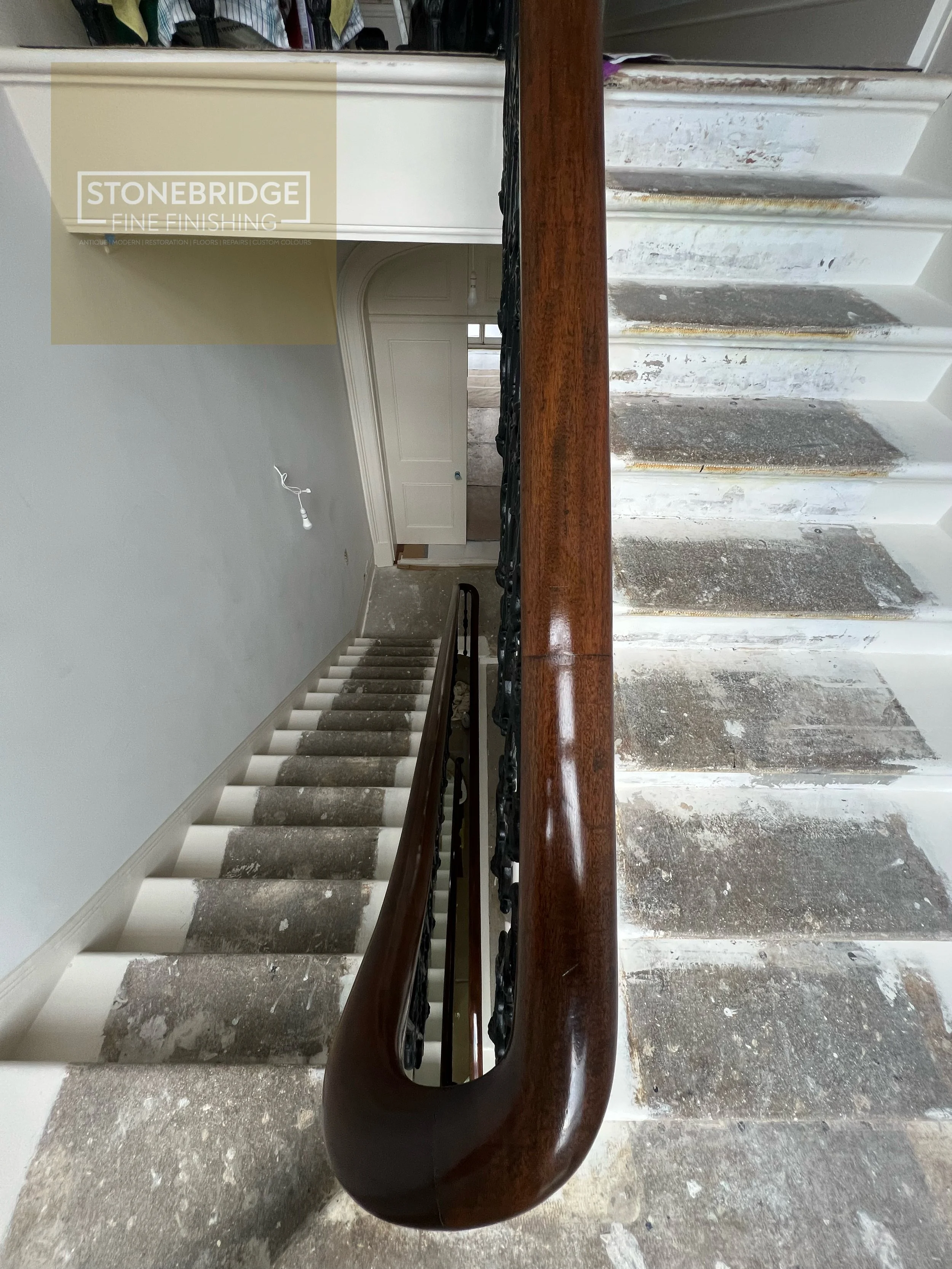 The image shows a staircase under renovation with a wooden handrail in the foreground, stairs covered in dust and debris, and a door visible at the top of the stairs.
