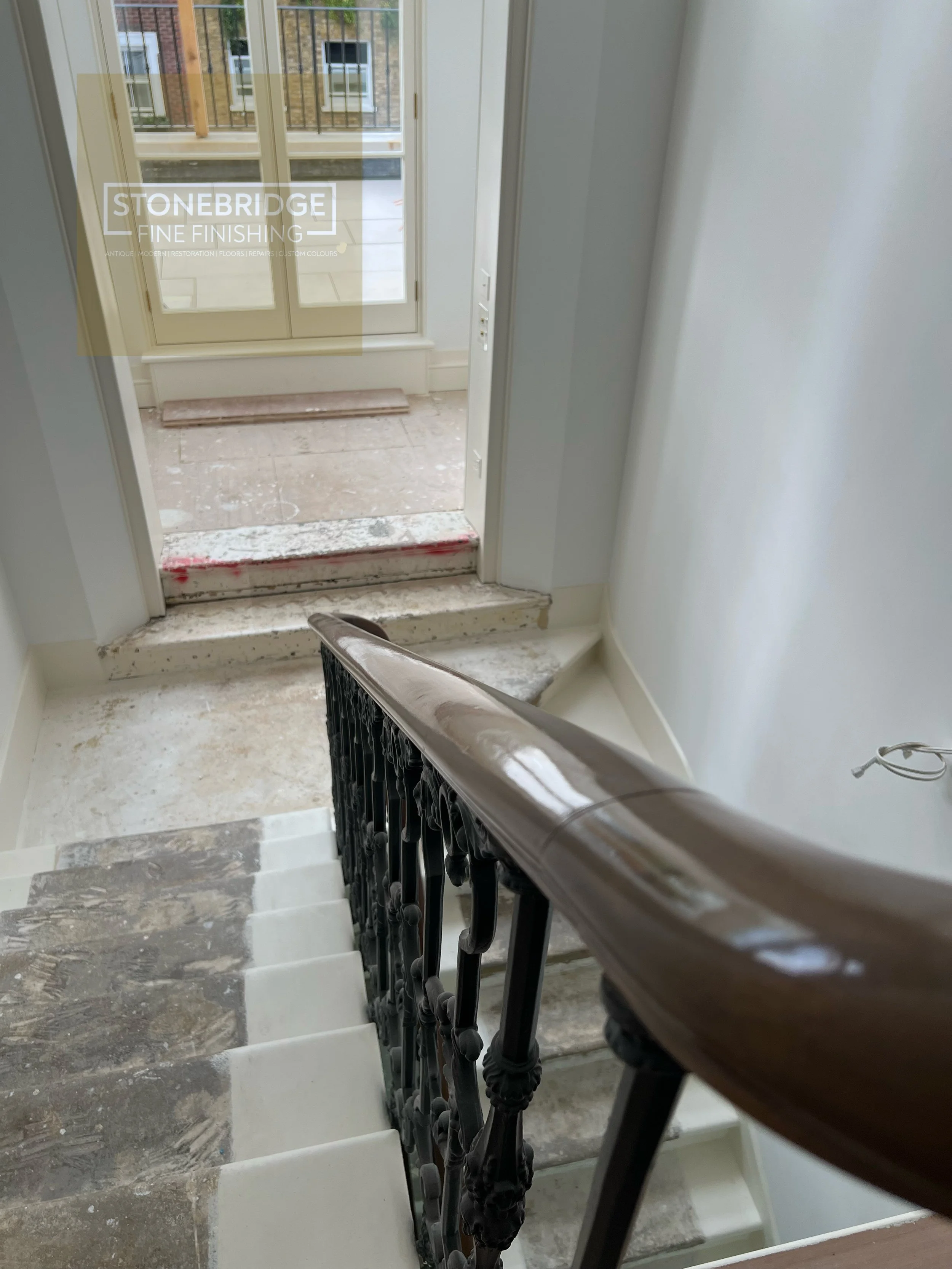 View of a stairwell with unfinished stairs, a black decorative railing, white walls, and a door leading outside, with construction or renovation work in progress.