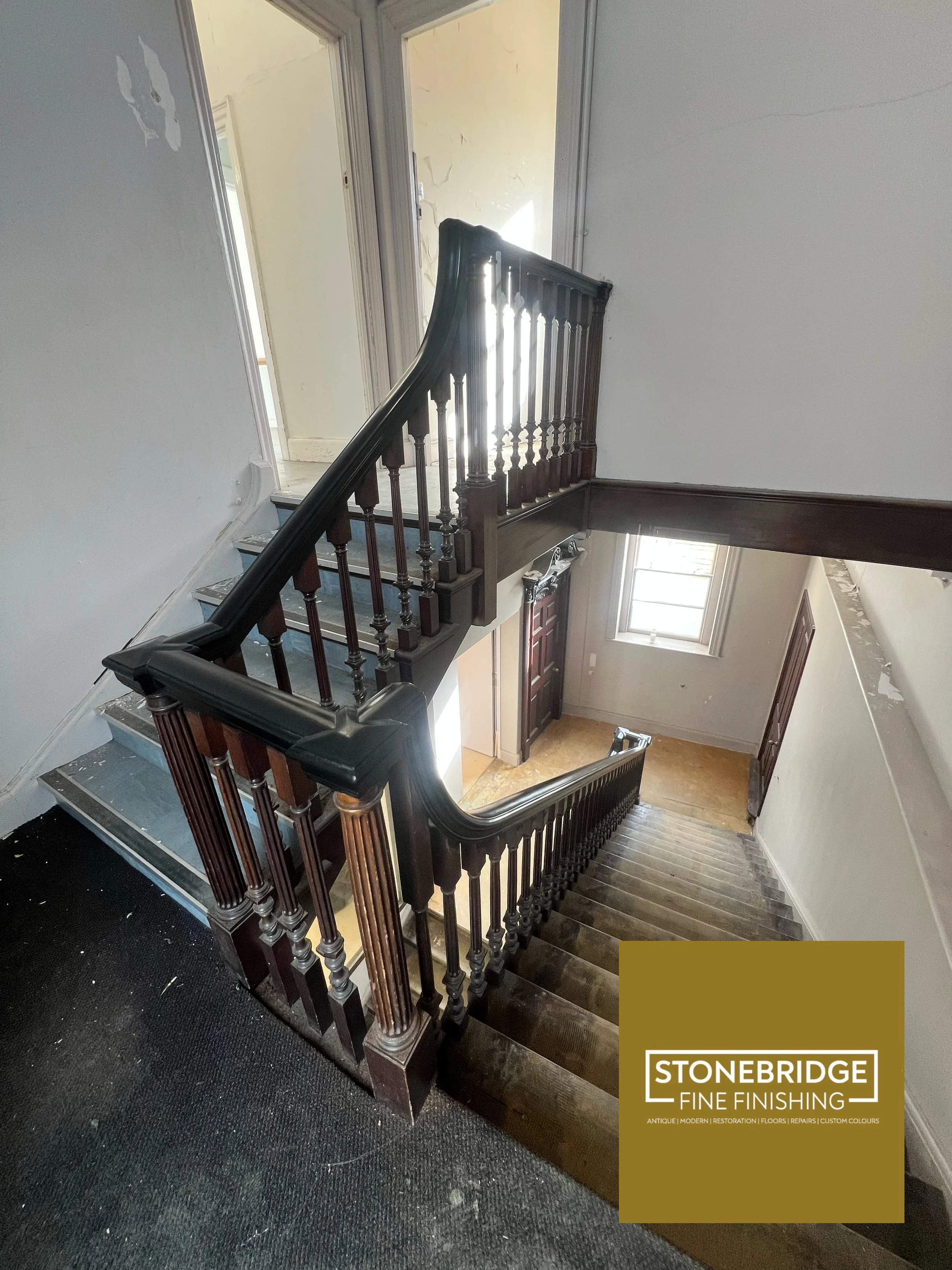 Interior view of a staircase with wooden banisters and steps. The staircase descends to a door below. The walls are worn with peeling paint, and there's a window at the bottom letting in natural light. A black carpeted area covers part of the stairca