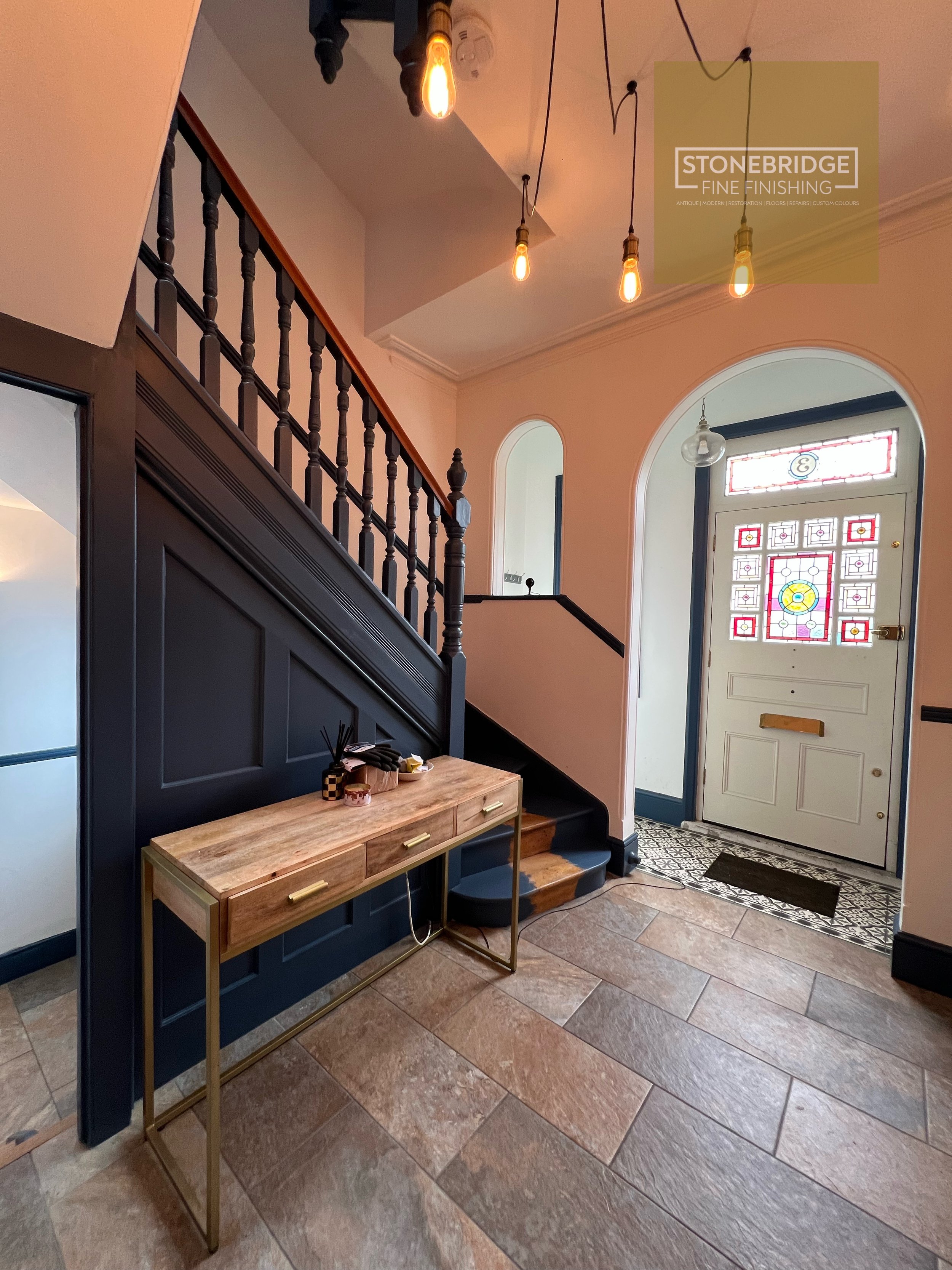 Interior view of an entryway with a dark blue wall panel, a small wooden table, and a decorative stained glass front door. Light fixtures with exposed bulbs hang from the ceiling, and there is a stairway with wooden handrail and black spindles.