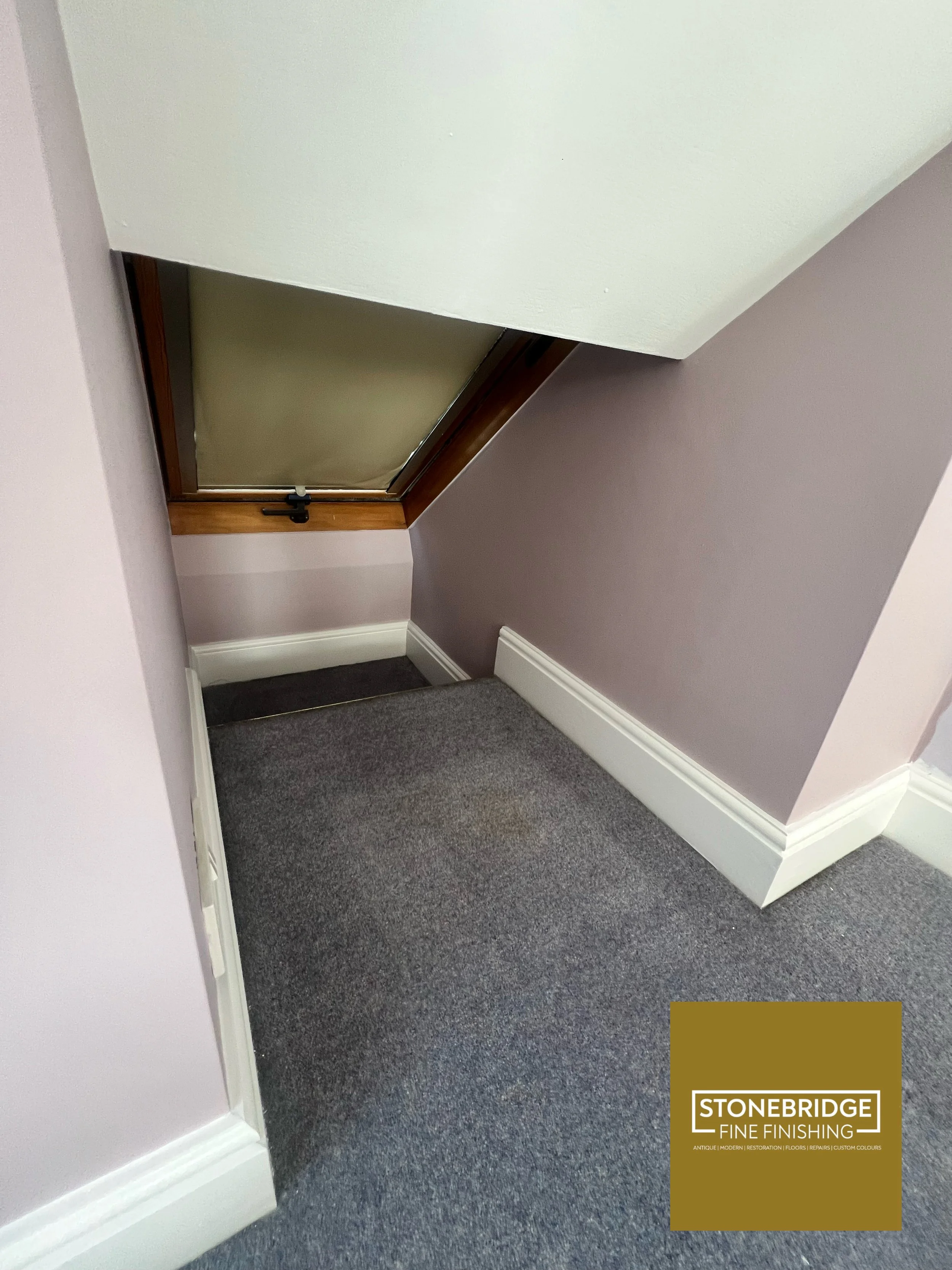 Interior corner of a room with a sloped ceiling, purple and white walls, a gray carpeted floor, and a small door with a black latch. There is a Stonebridge Fine Finishing logo in the bottom right corner.