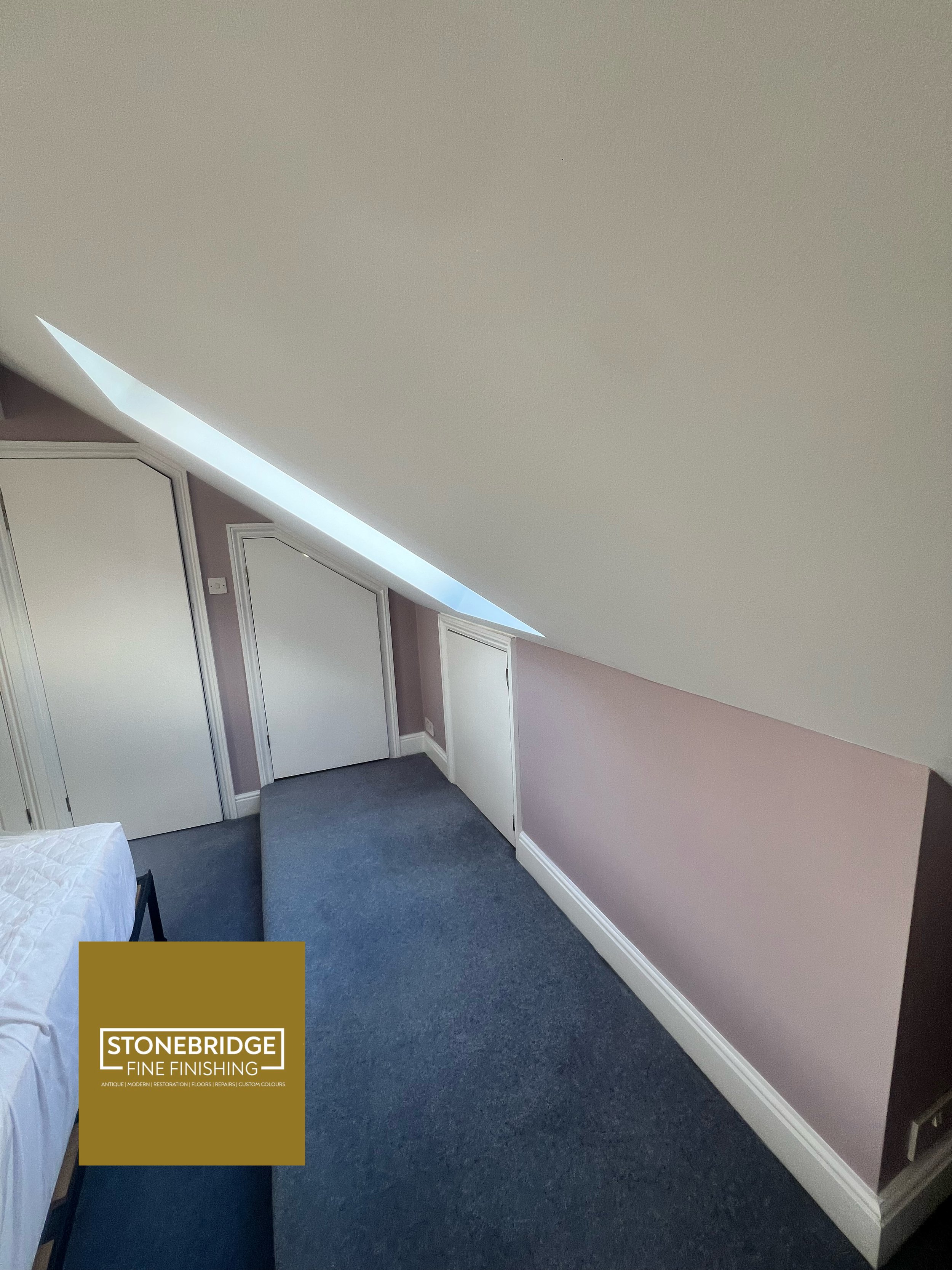 A room with sloped ceiling, two closed white doors, a small window, and a purple wall with white baseboard. Part of a bed with white bedding and a black chair are visible. There is a brown and white logo in the bottom left corner reading 'STONEBRIDGE