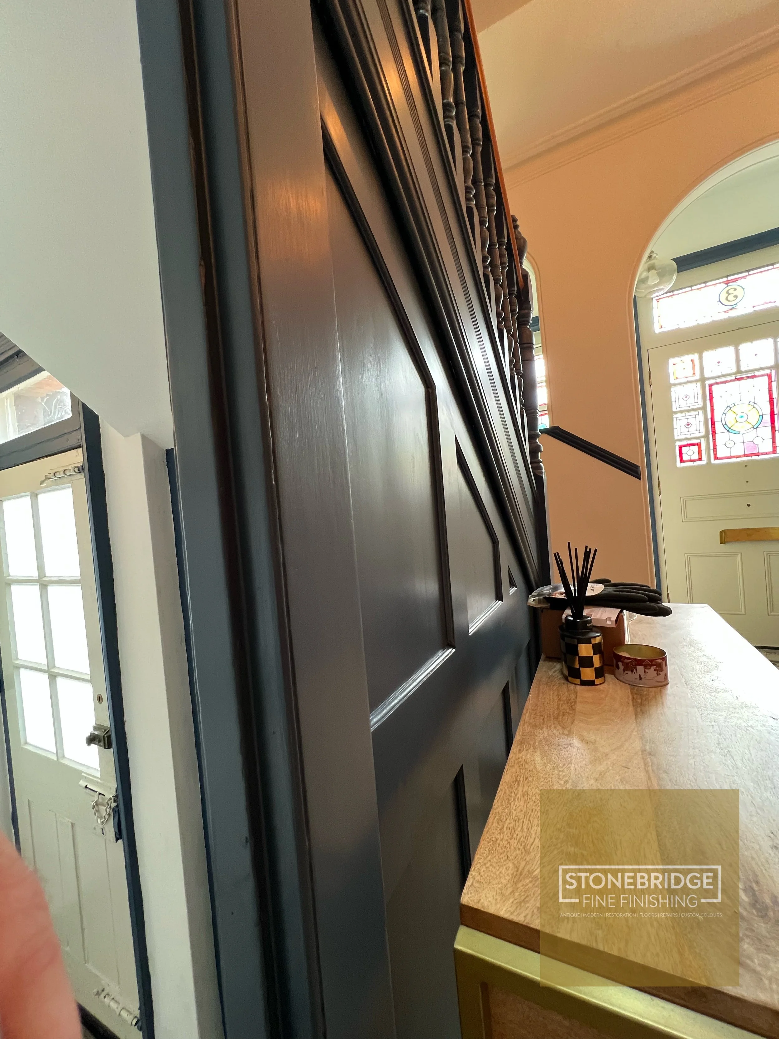 Close-up view of a dark wooden cabinet, possibly in a kitchen or dining area, with a countertop next to it. The background includes a door with stained glass and some decorative items on the counter, like reed diffusers and a small container. The spa