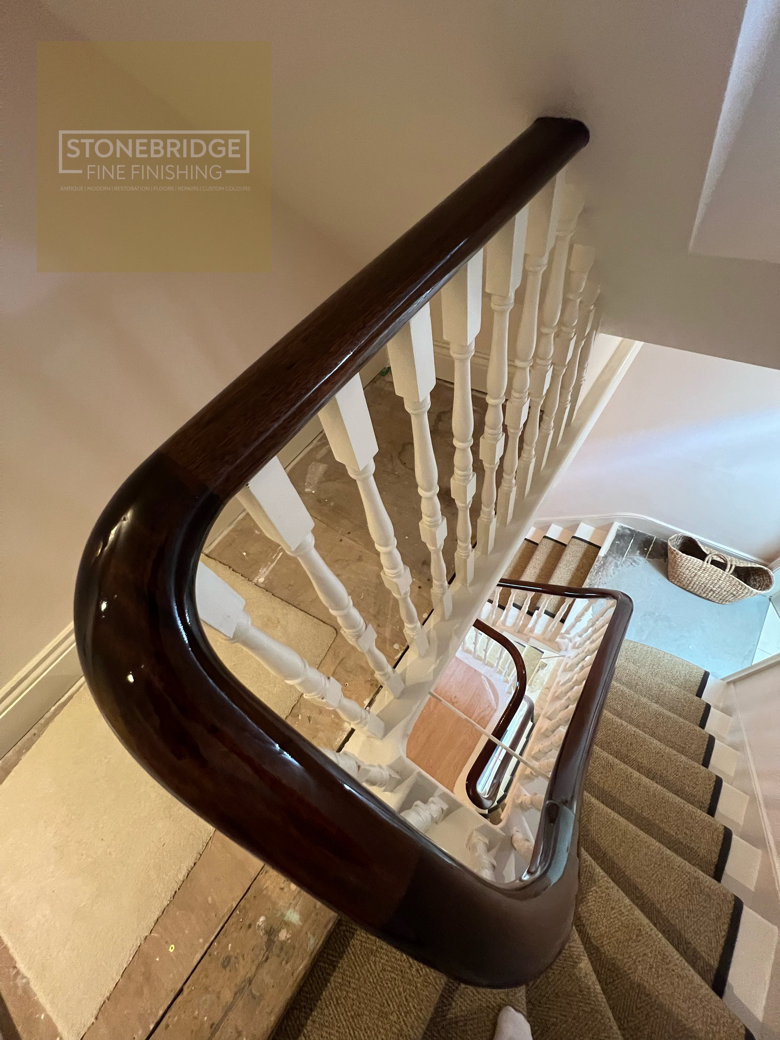 Top view of a staircase with a dark wooden handrail, white spindles, beige carpet, and cardboard flooring at the bottom. A basket is visible on the lower floor.