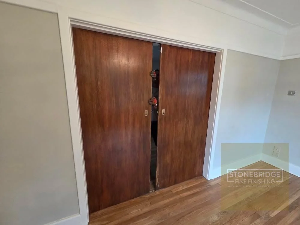 Wooden sliding closet doors partially open, showing a person taking a photo in a room with hardwood flooring and light-colored walls.