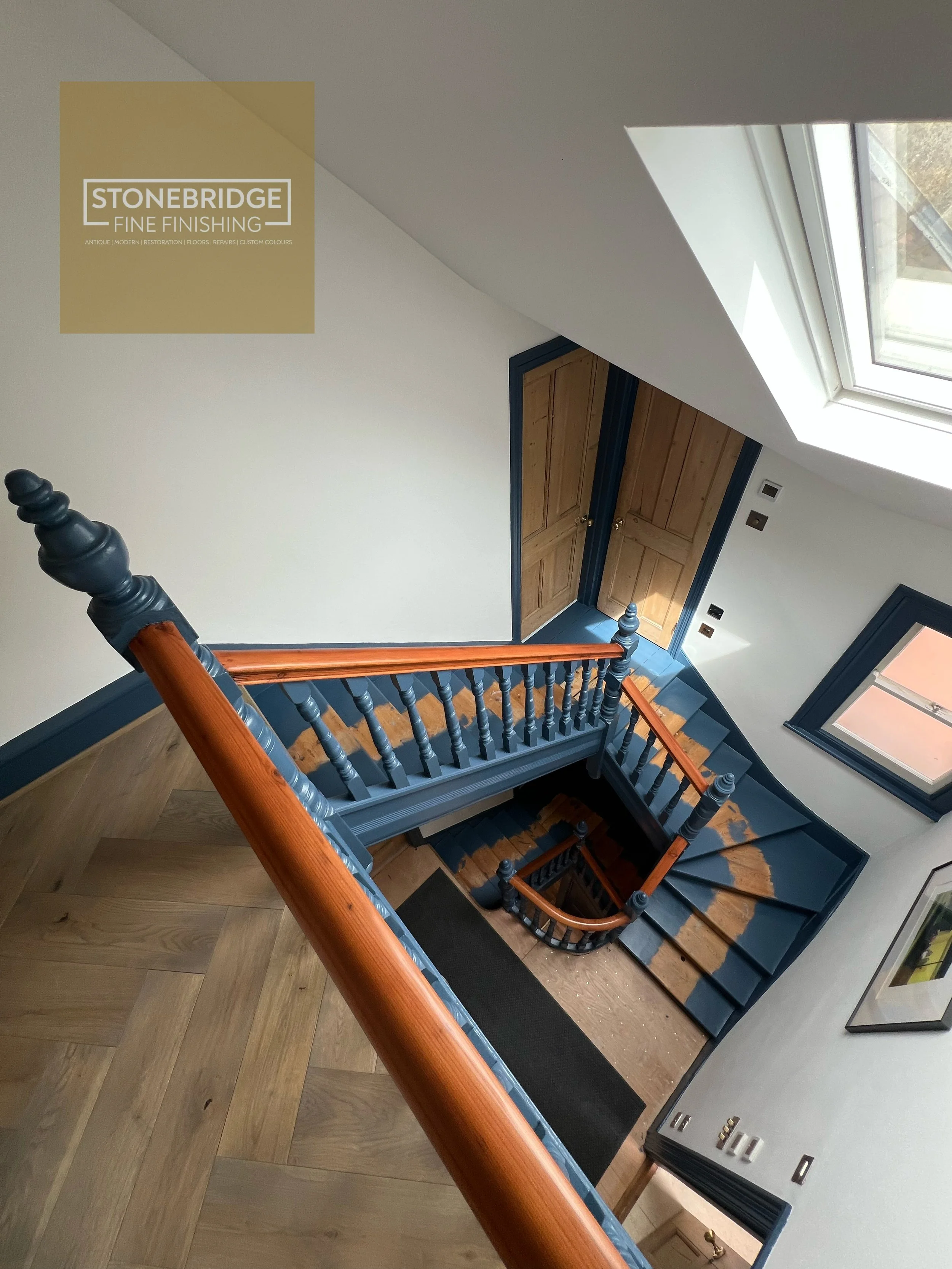 View of a staircase with blue and wood-colored handrails, wooden stairs, a white wall, a large window letting in natural light, and a door with wooden panels at the bottom of the stairs.