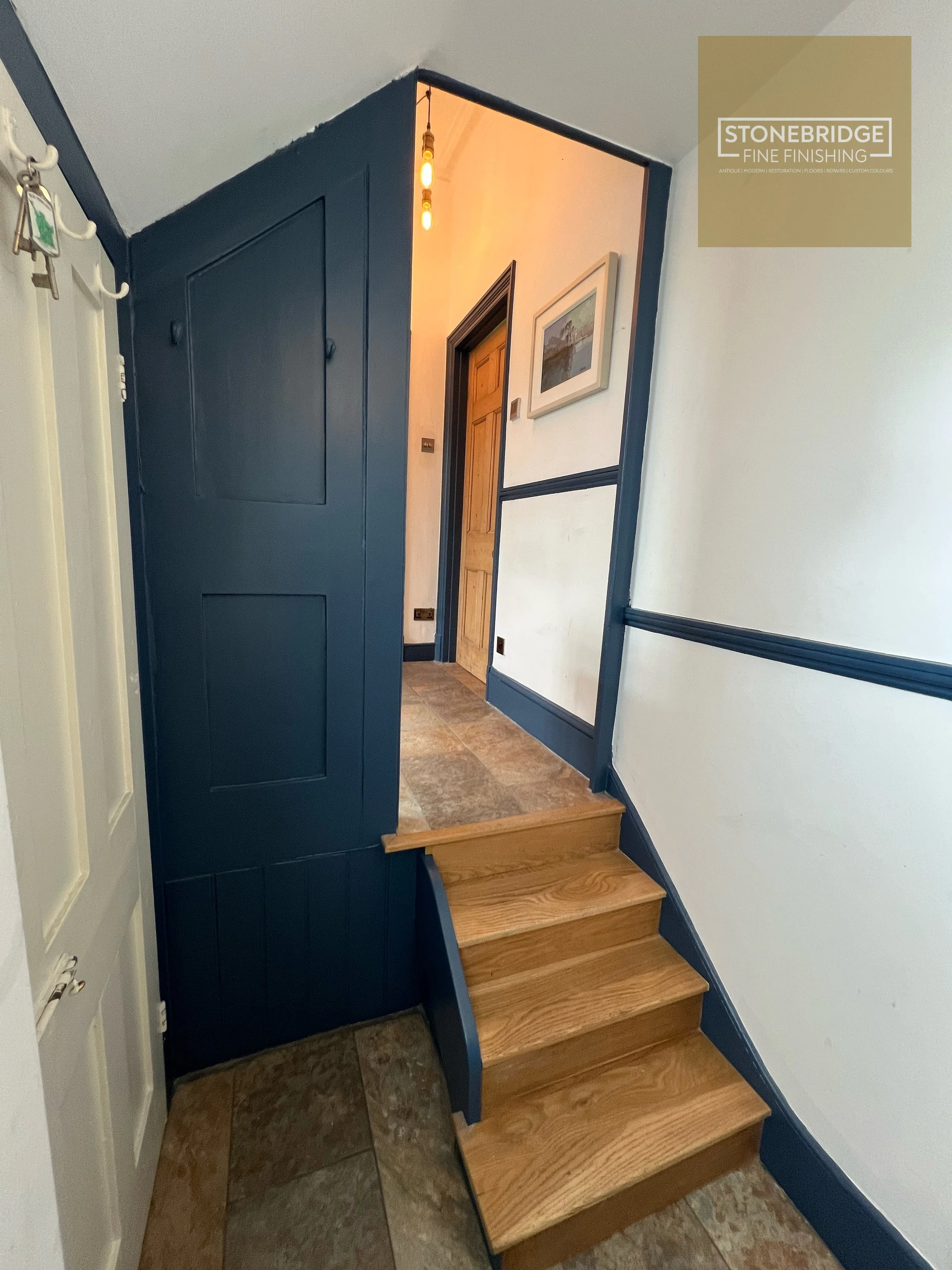 Interior view of a staircase with four wooden steps, a dark blue trim, and white walls with black trim. There is a framed picture on the wall, a small step, and a corridor with a wooden door. The ceiling has a hanging bulb light. A closet door is vis