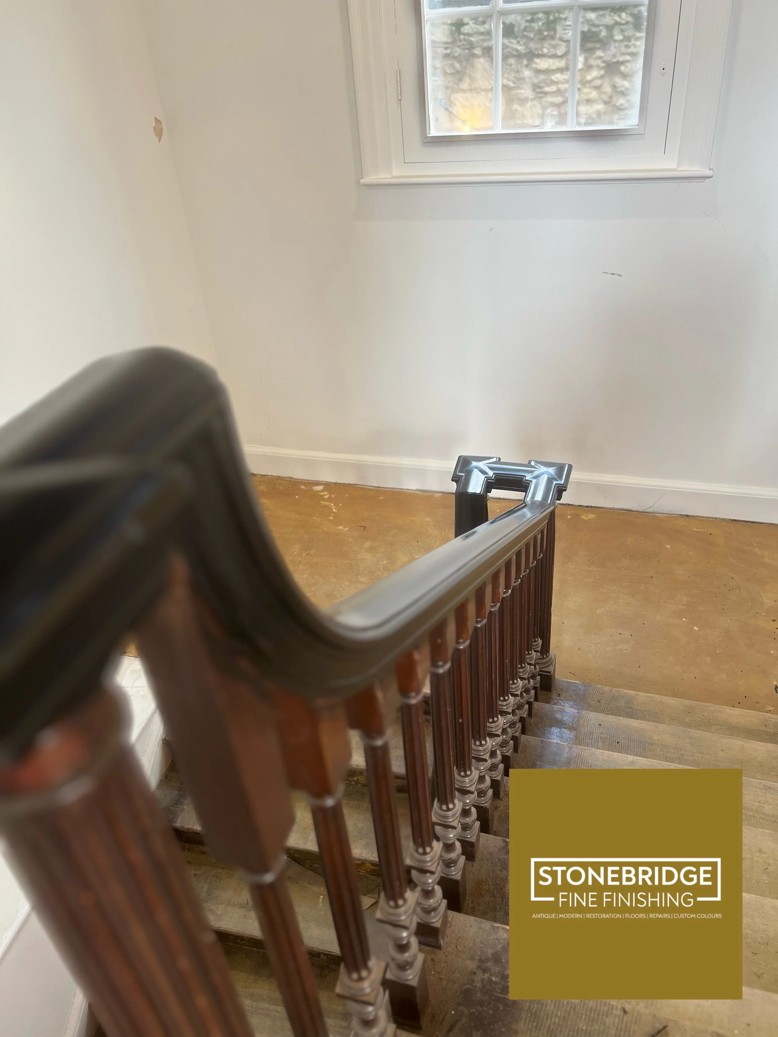View of a staircase with wooden handrails and brown spindles, leading down to a wooden floor. A window with a stone wall outside is visible at the top of the stairs. There is a logo in the bottom right corner that reads "STONEBRIDGE FINE FINISHING" w