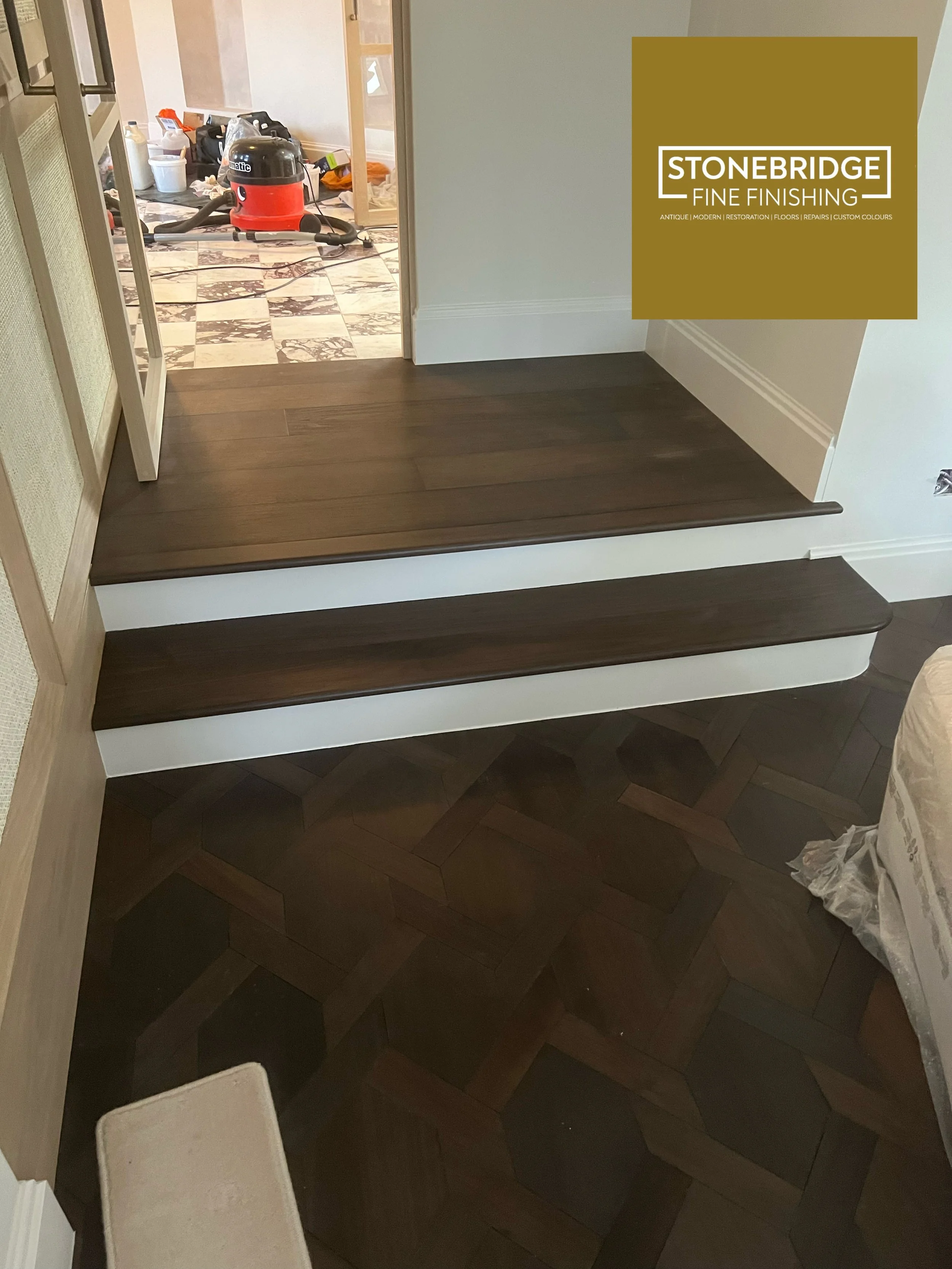 Image shows a room with dark wooden flooring and steps leading to a room with marble flooring. There is a vacuum cleaner in the background and some scattered items on the marble floor.
