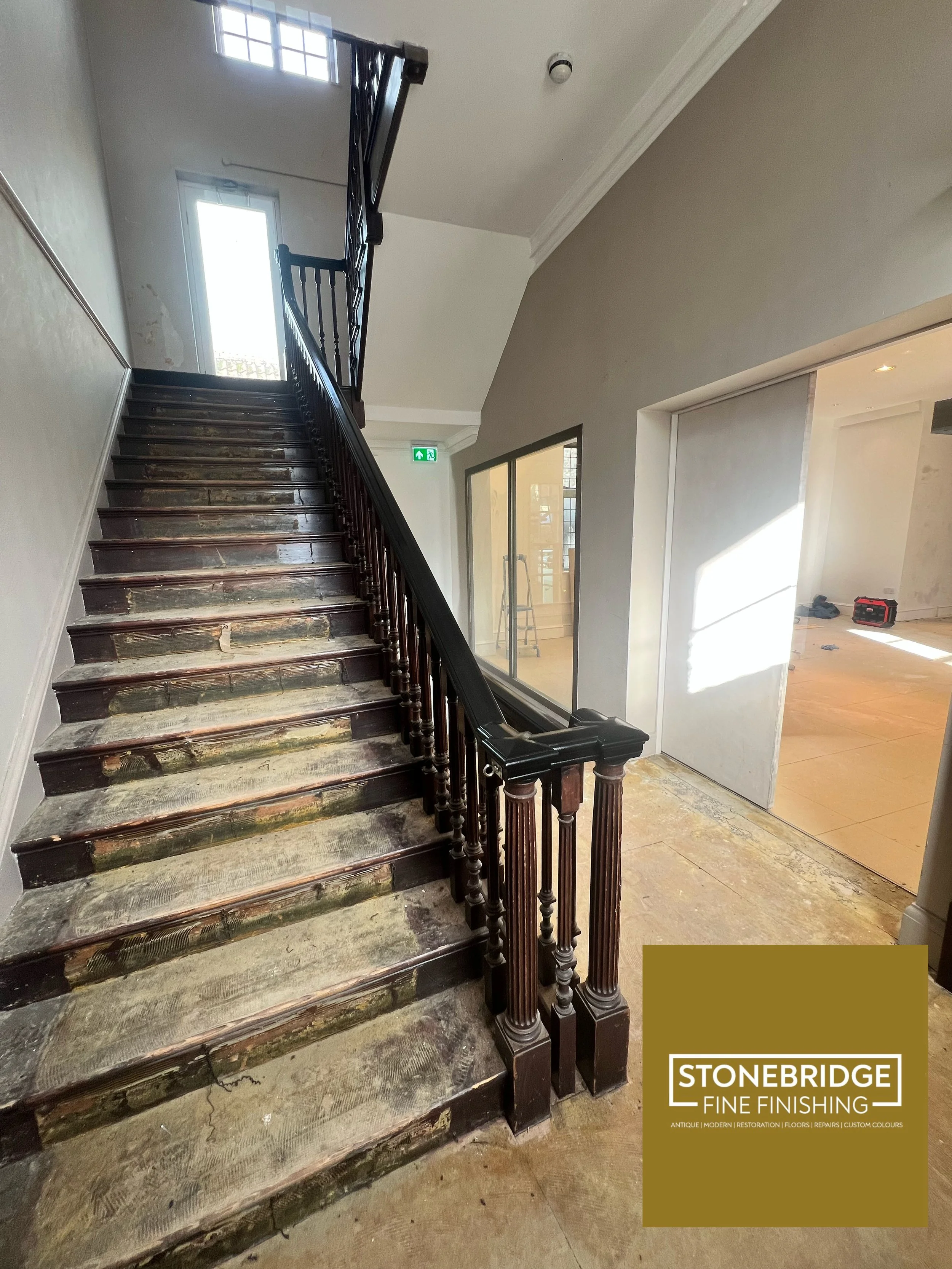 Interior staircase with wood steps showing signs of wear and tear, black wooden handrail and balusters, adjacent glass door and window, existing construction or renovation materials, and a partially completed wall with tools.