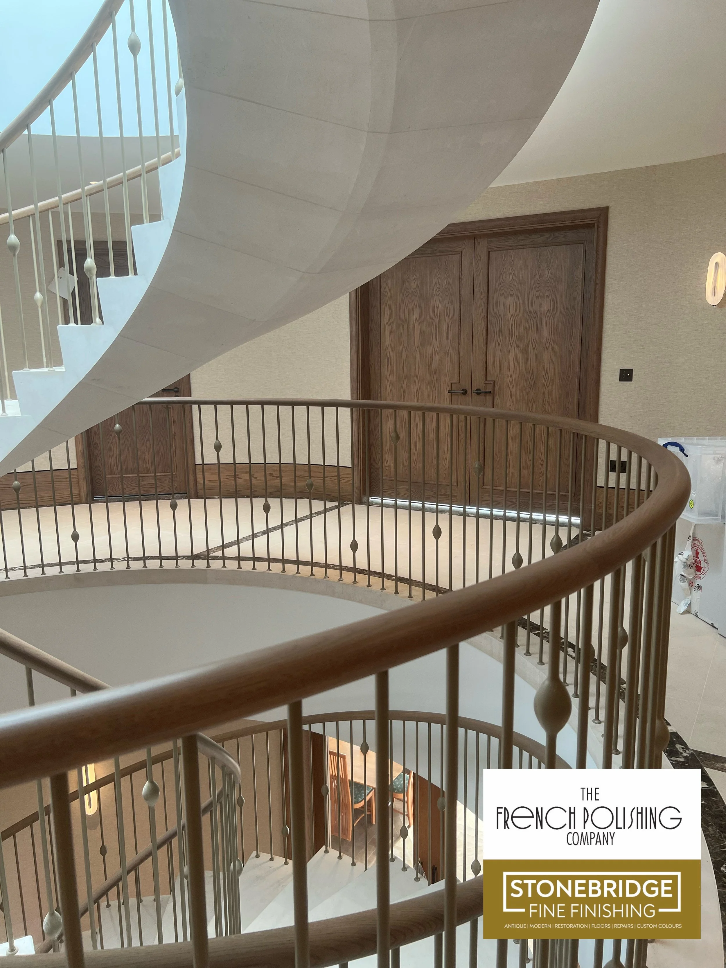 Interior view of a staircase with wooden handrails and metal balusters, overlooking a lower level with chairs and a table. A closed wooden door is in the background, with a wall-mounted decorative object on the right.