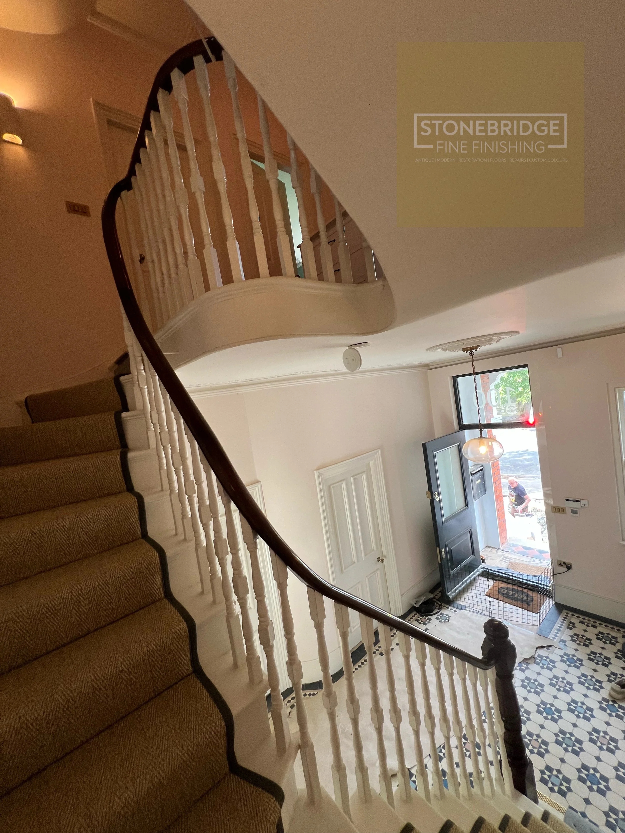 Interior view of a house entrance with a brown carpeted staircase to the left, white balustrades, and an open front door leading outside. There is a pendant light hanging from the ceiling and a window with trees visible outside.