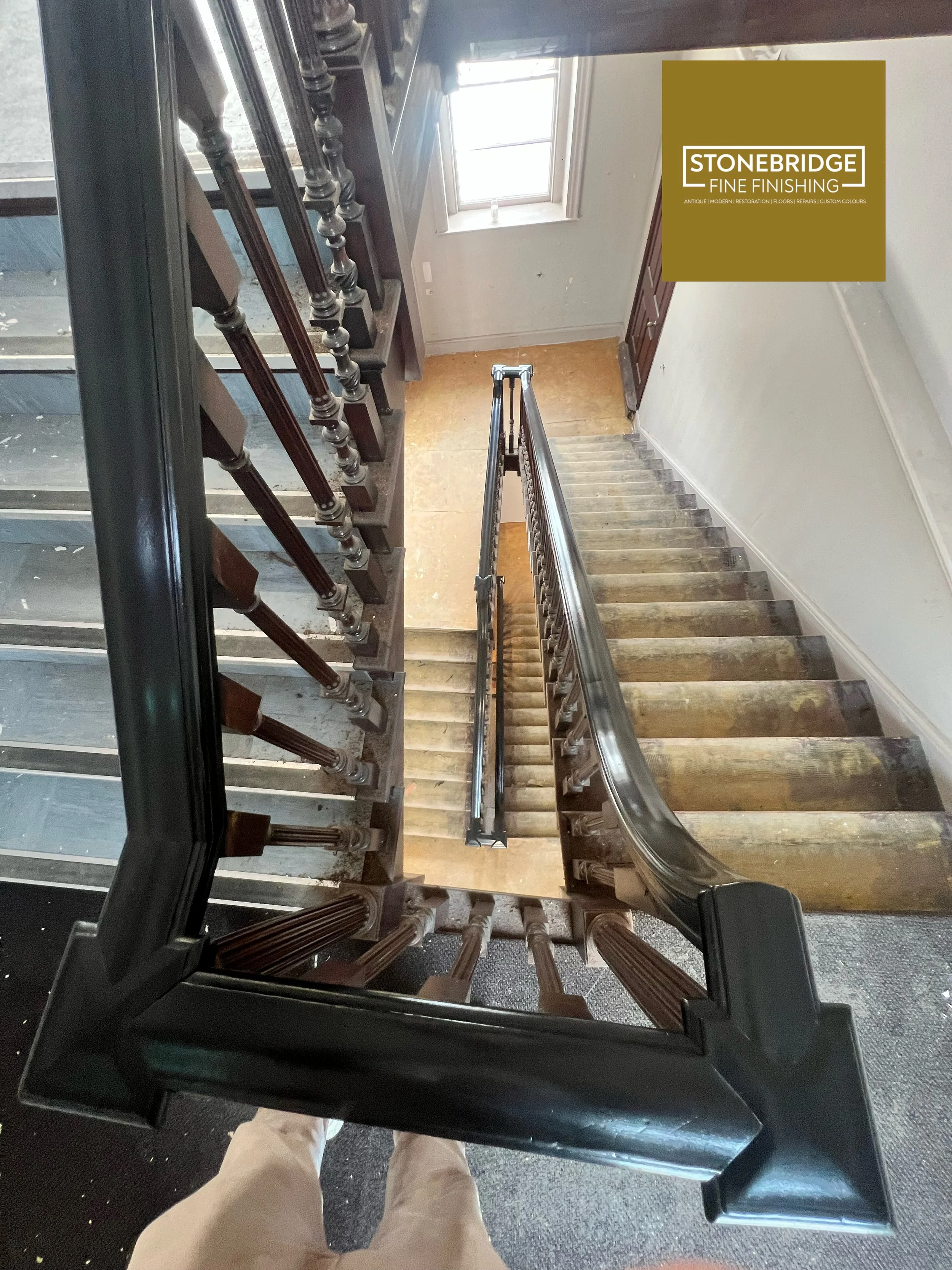 A view looking down a staircase with wooden banisters and a metal railing, taken from the top of the stairs. At the bottom, a partially visible person wearing khaki pants and light-colored shoes is standing on a carpeted floor. The wall on the right 