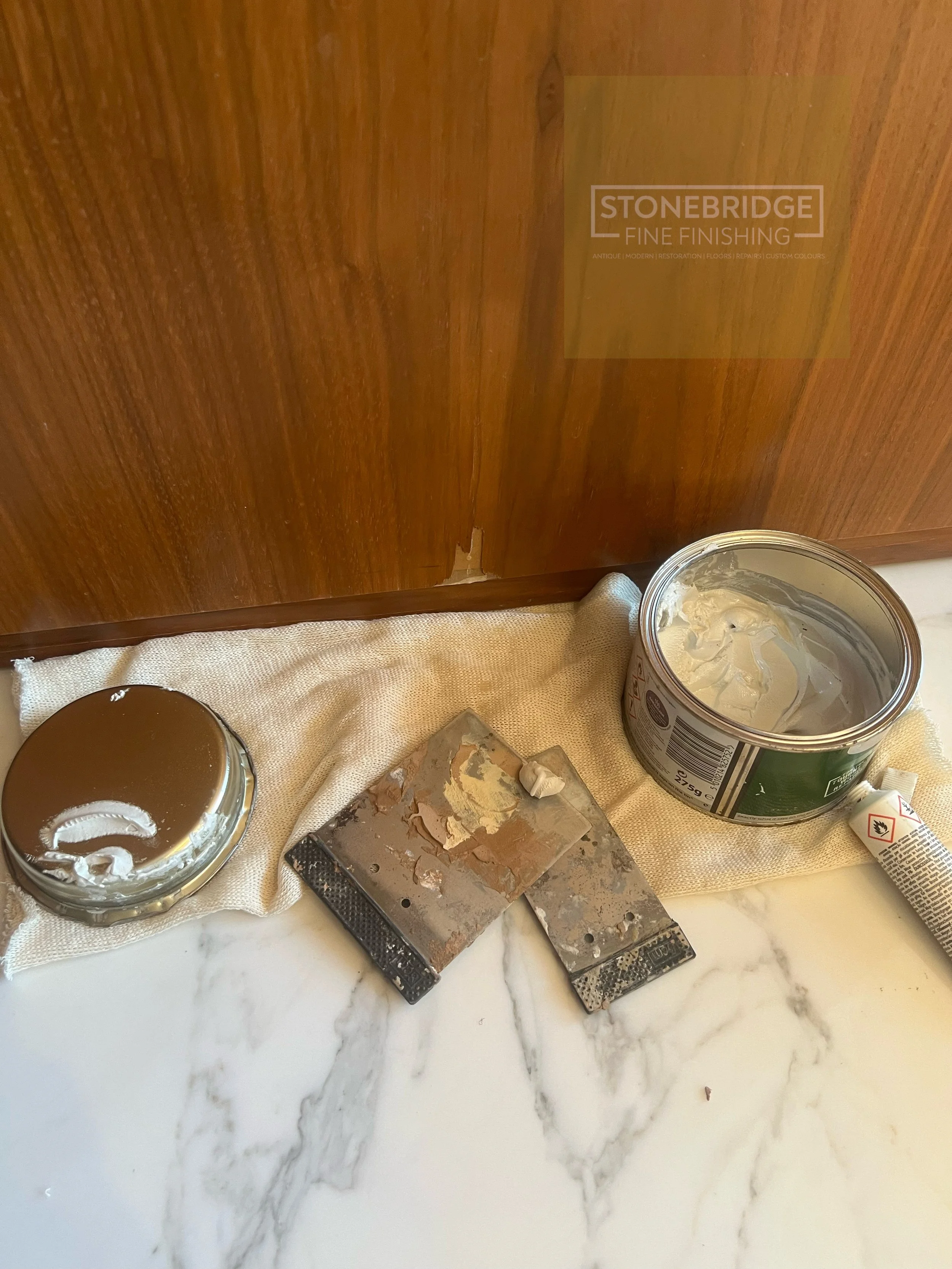 A marble countertop holding a can of cream, a black spatula, and a small container of brown paint, with a wooden surface and a paint can in the background.