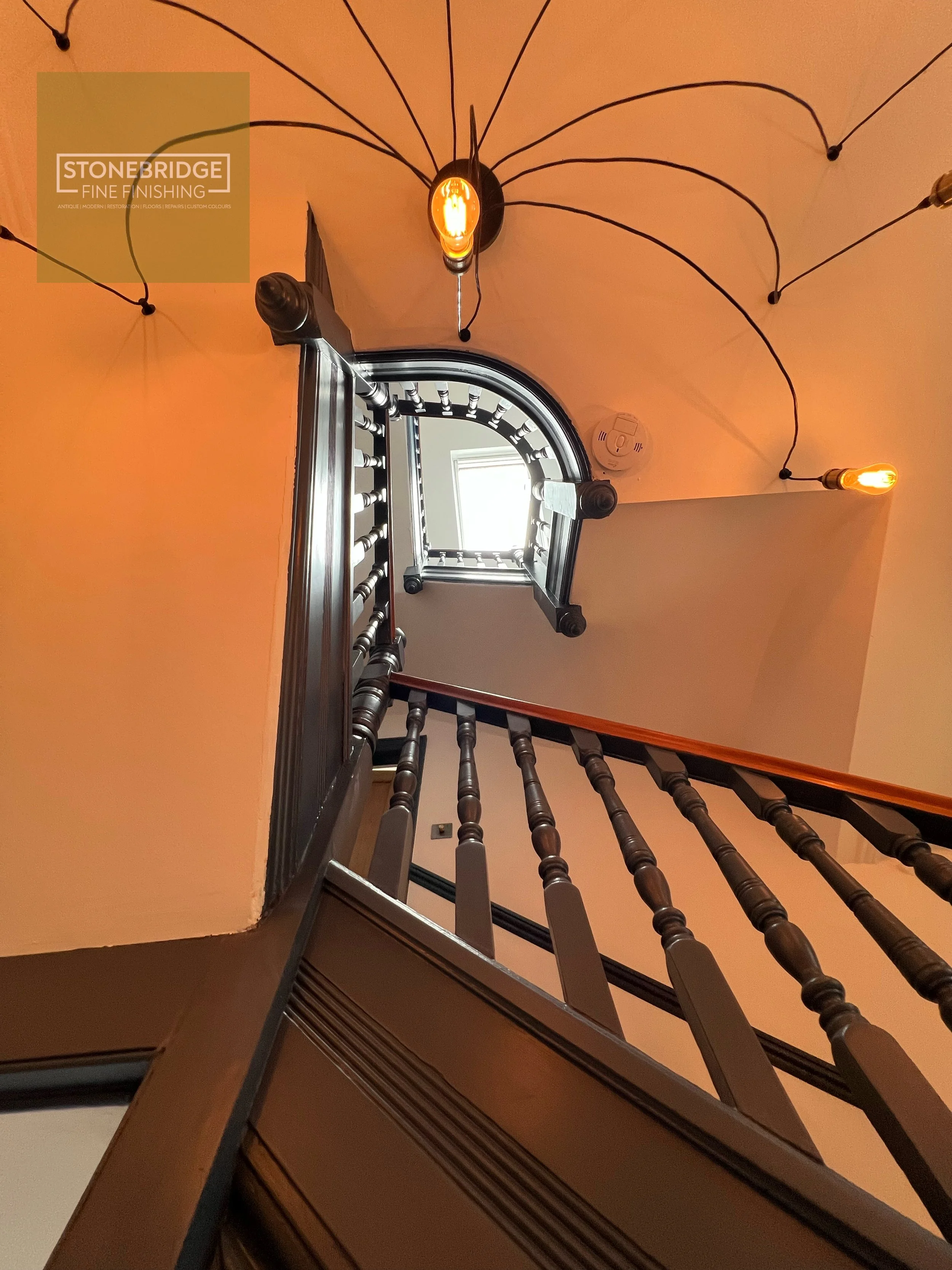 A view upwards from the bottom of a staircase with dark wooden banisters and spindles, looking towards a small arched window at the top and wall-mounted lighting fixtures with exposed filament bulbs on the ceiling.