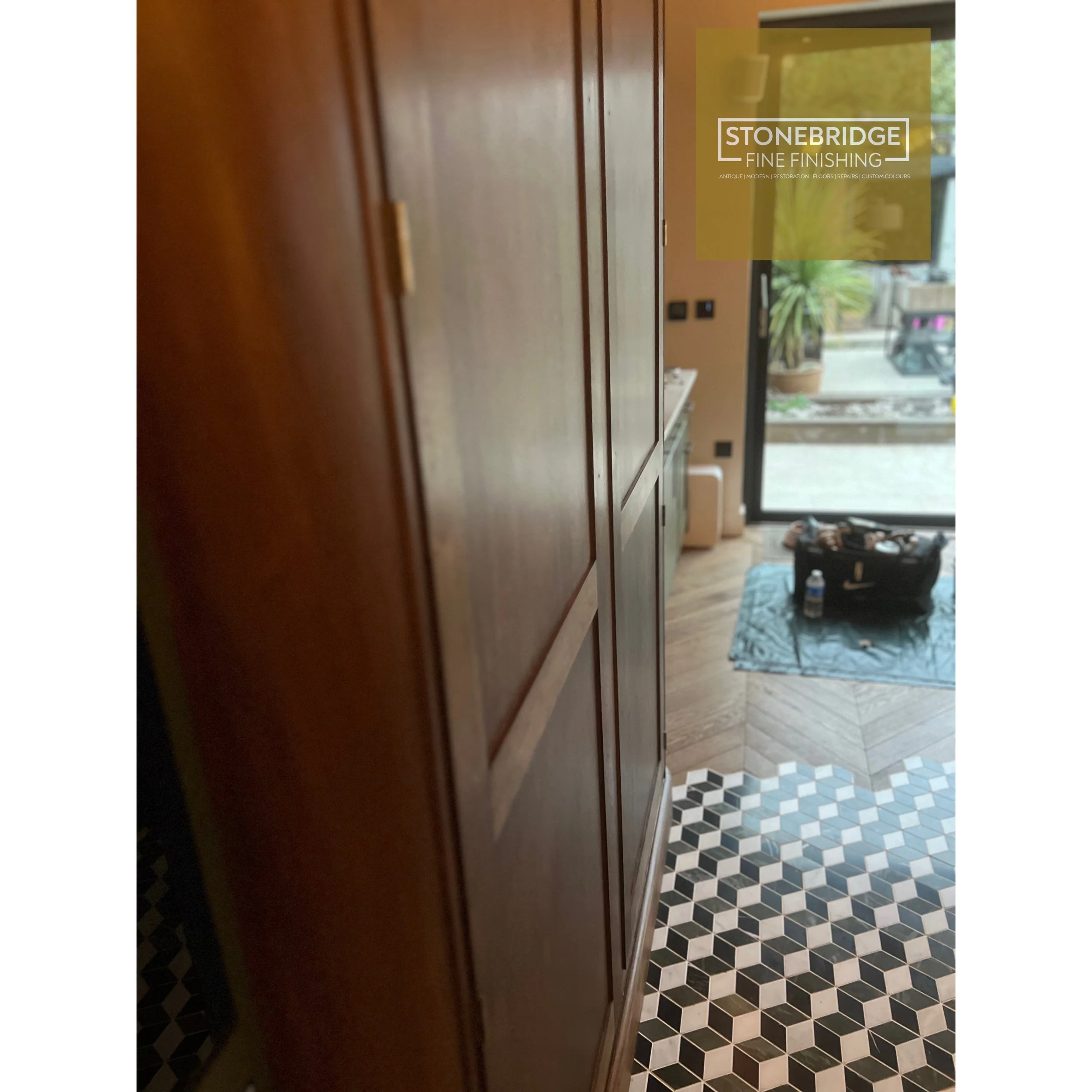 Wooden cabinet doors next to a floor with black, white, and gray geometric tiles. In the background, a doorway with glass panels shows a living area with plants, furniture, and a window.
