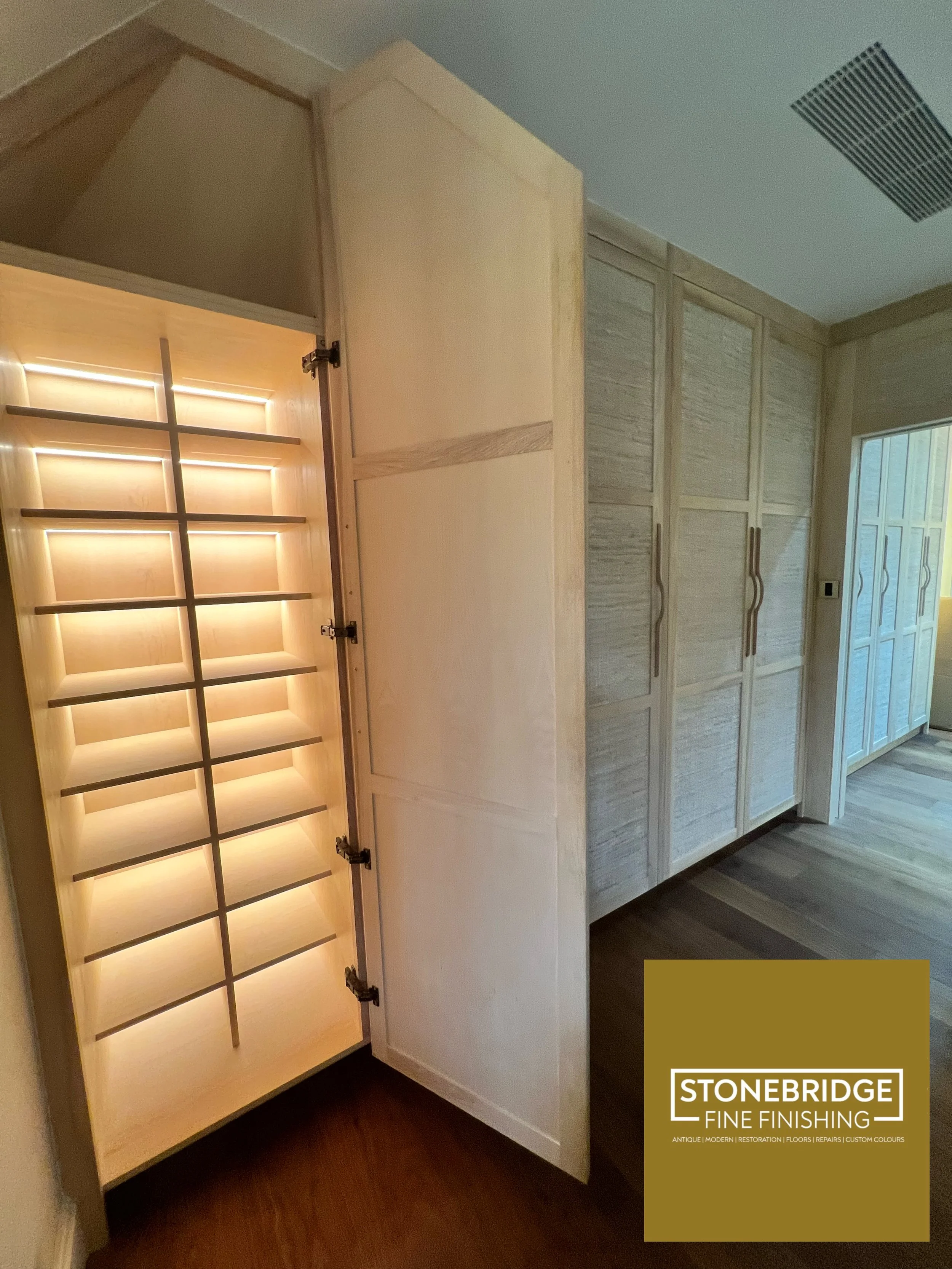 Interior view of custom built wooden cabinetry, with a gown display frame with shelves inside, and a row of cabinets with handles to the right, in a room with wood flooring and an air vent on the ceiling.