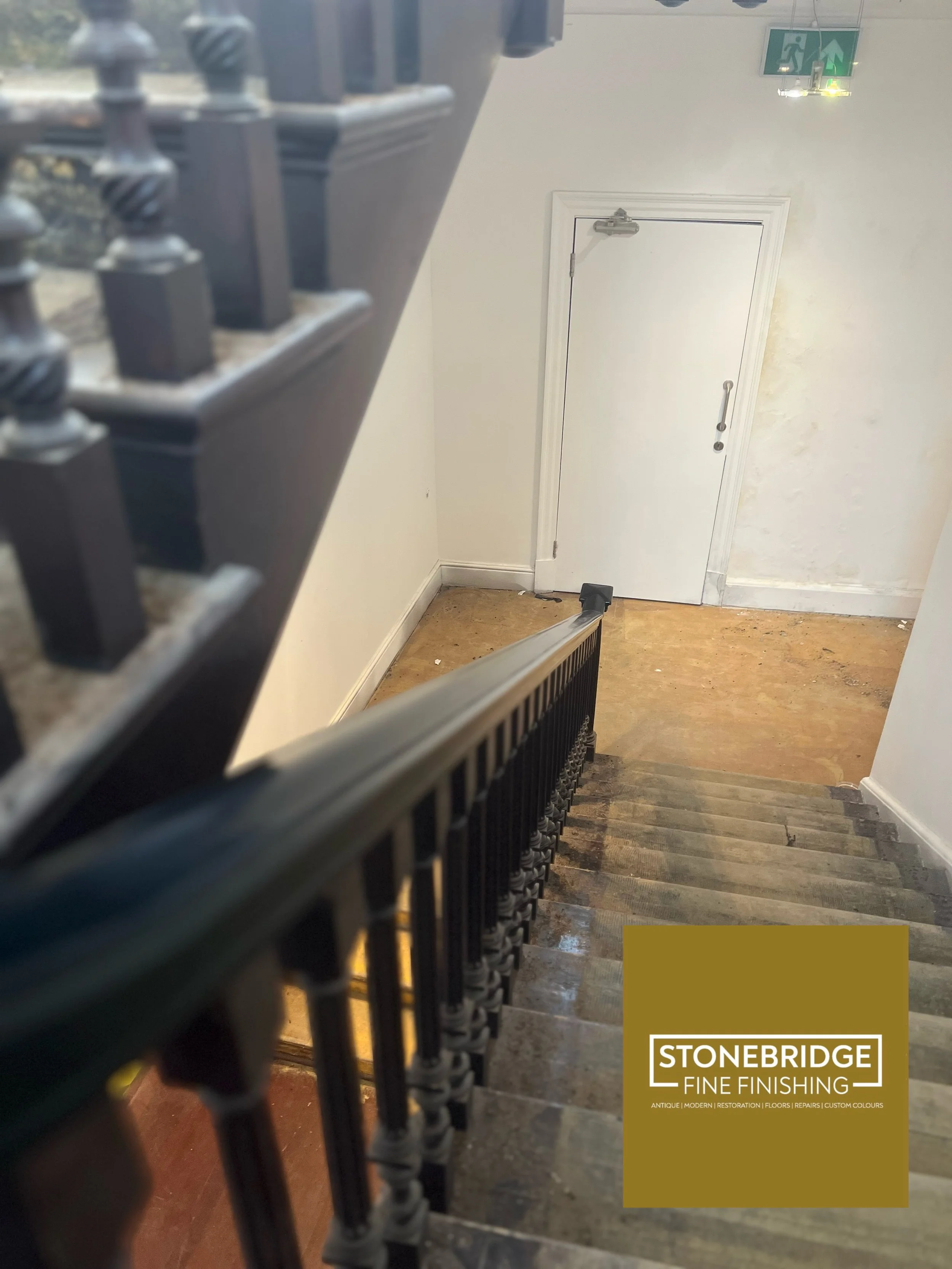 A staircase with a black metal railing leading down to a door at the bottom. The door is white with a handle and a door closer at the top. There is a green exit sign with a running figure and an arrow pointing upward hanging from the ceiling above th