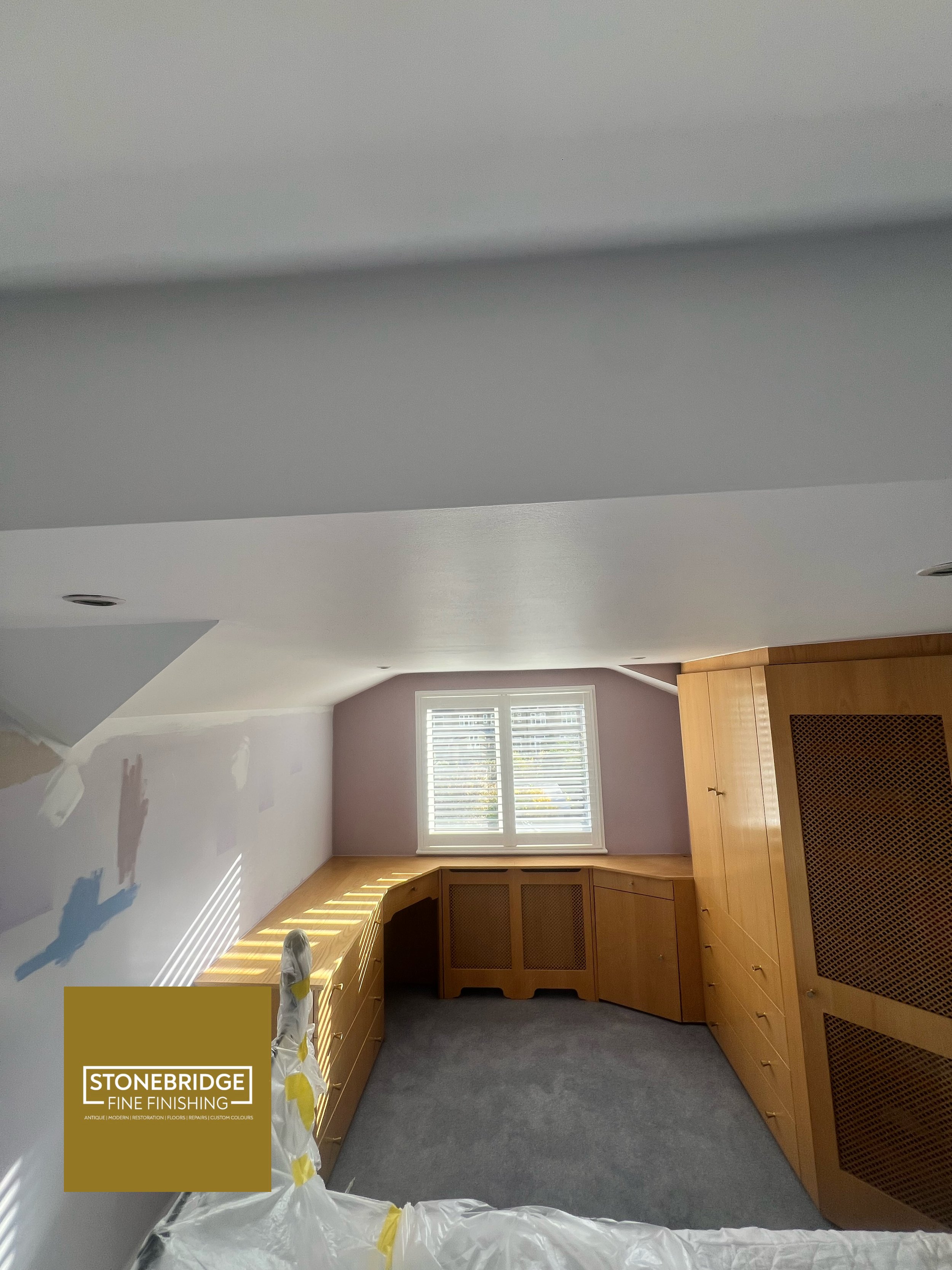 Photo of a room with white walls, a window with blinds, wooden cabinets, and a gray carpeted floor during renovation or remodeling, with a partially painted wall.