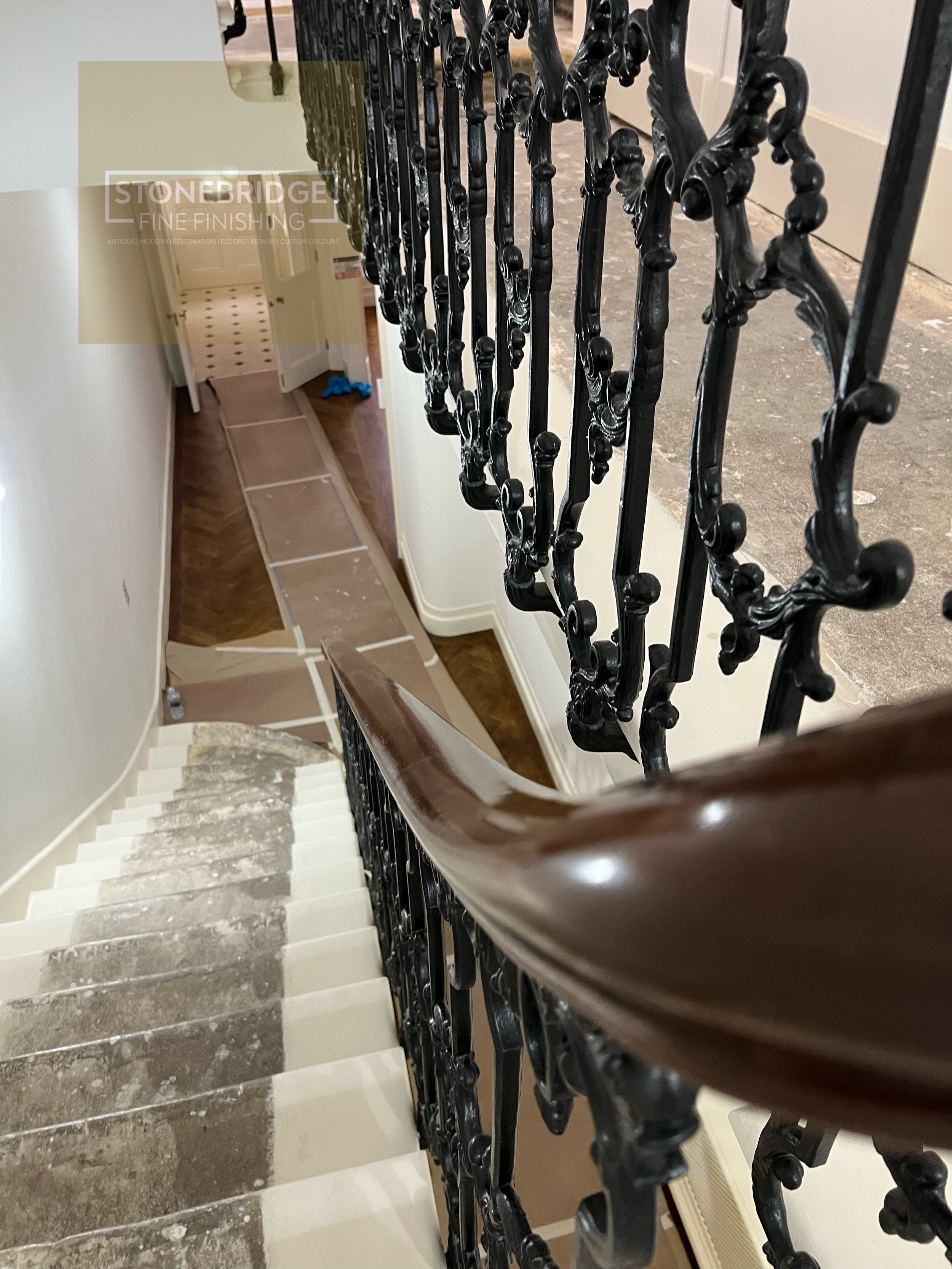 View of a staircase with a decorative iron railing and a wooden handrail, looking down from the top of the stairs. The staircase has white risers and dark treads, with some areas covered in masking tape, indicating ongoing renovation work. The backgr